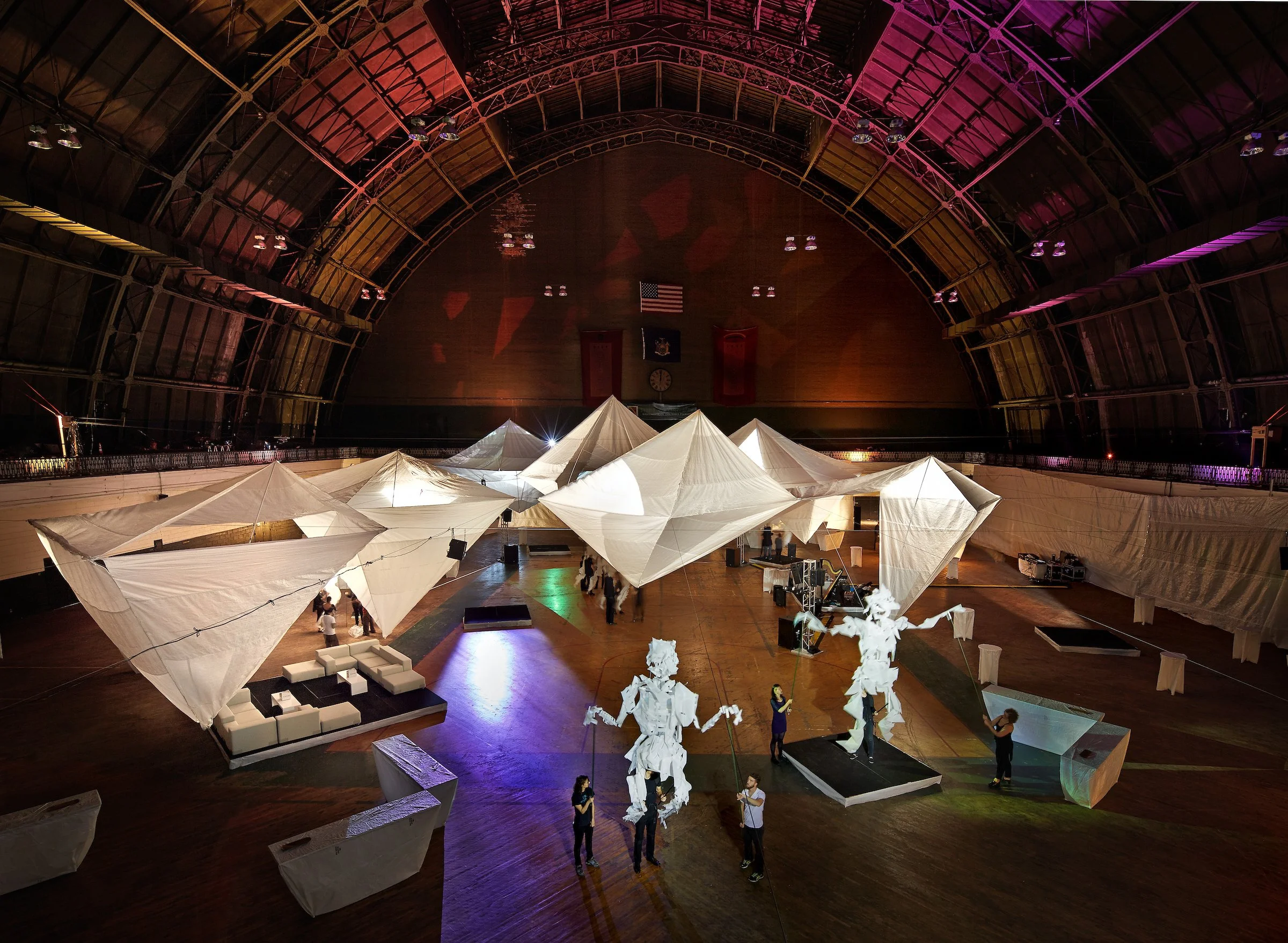 Event hall with white fabric tents, sculptures, and people setting up or preparing for an event in a large, curved ceiling space.