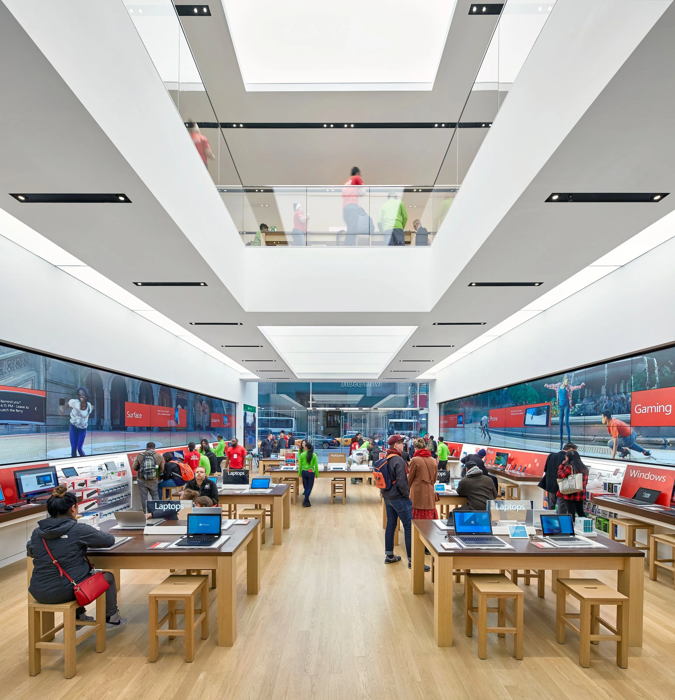 Inside a modern electronics store with tables displaying laptops and other devices, multiple customers shopping, LED advertisement displays along the walls, and people walking both inside and on the upper level.