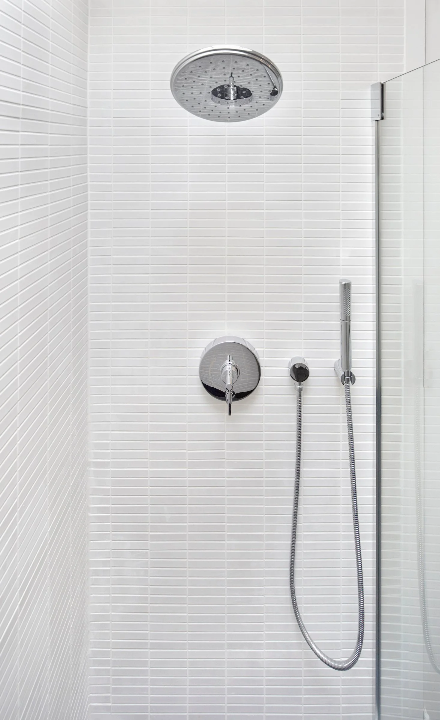 Minimalist white-tiled shower with a large round rain shower head, a control knob, and a handheld shower head with hose.