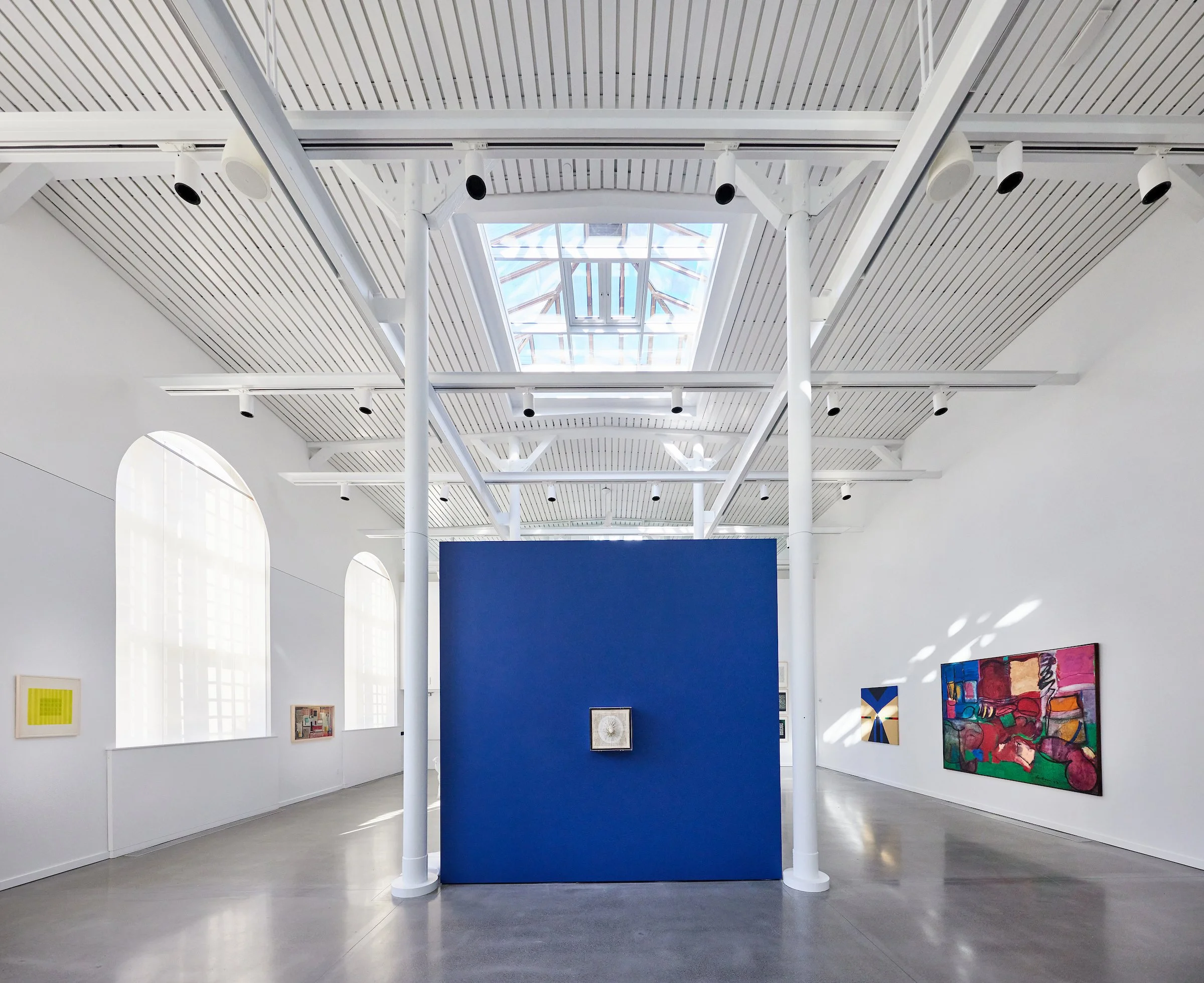 Modern art gallery with white walls, large arched windows, artworks on the walls, and a central blue partition with a framed piece of art. The ceiling features a skylight allowing natural light, with white structural supports and black spotlights.