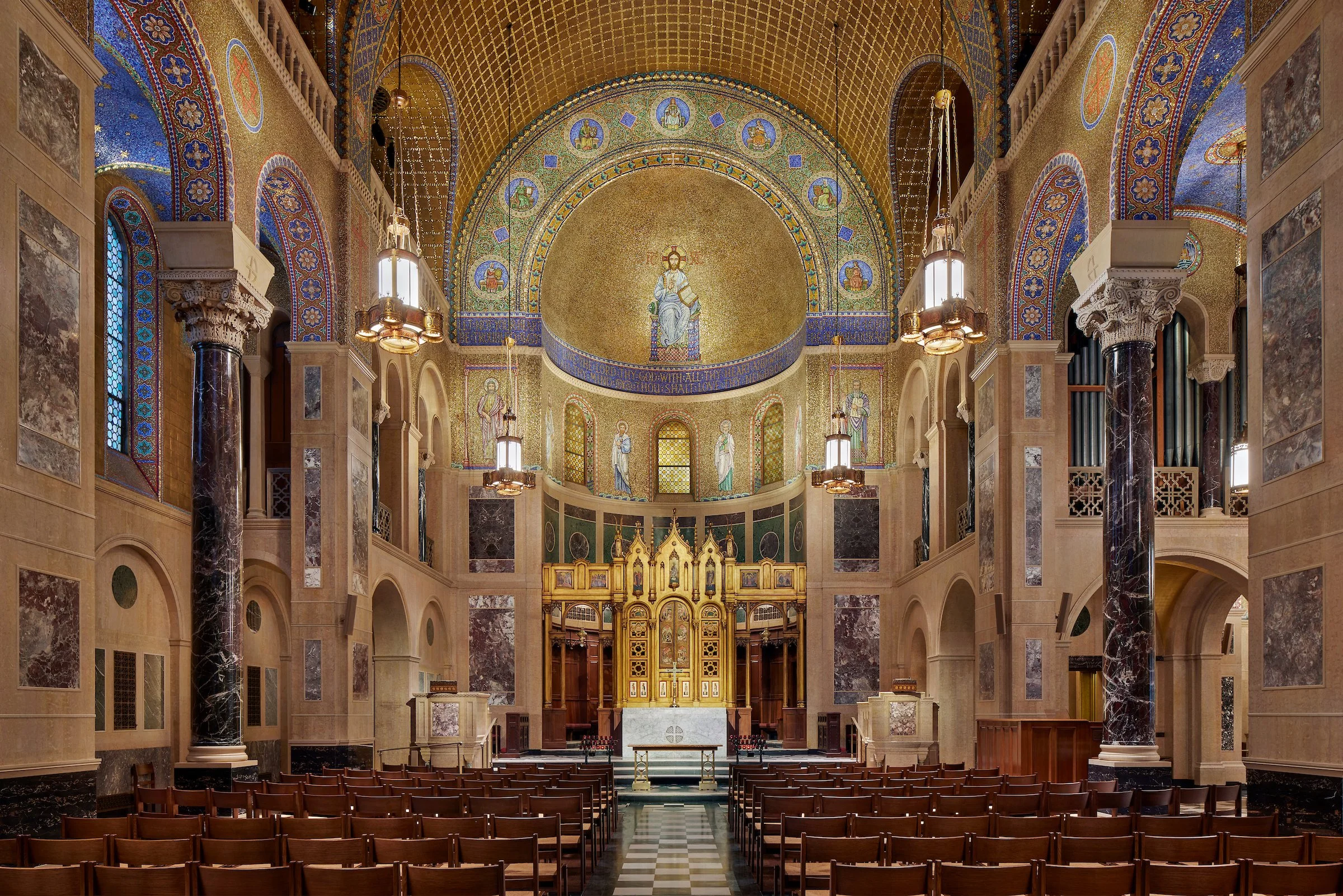 Interior of a richly decorated church with golden mosaics, tall marble columns, wooden pews, and stained glass windows.