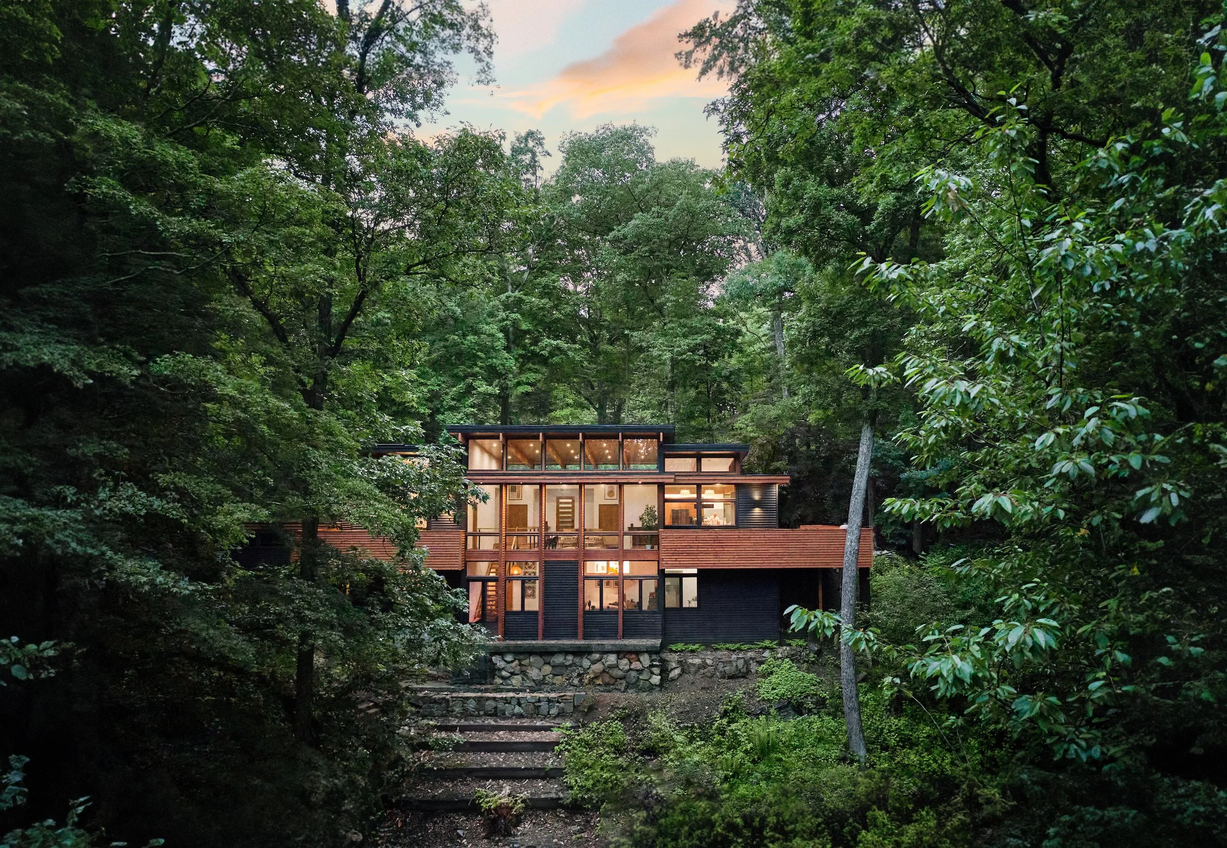 A modern house with large glass windows is built into a wooded hillside, surrounded by tall green trees, with a stone staircase leading up to it.