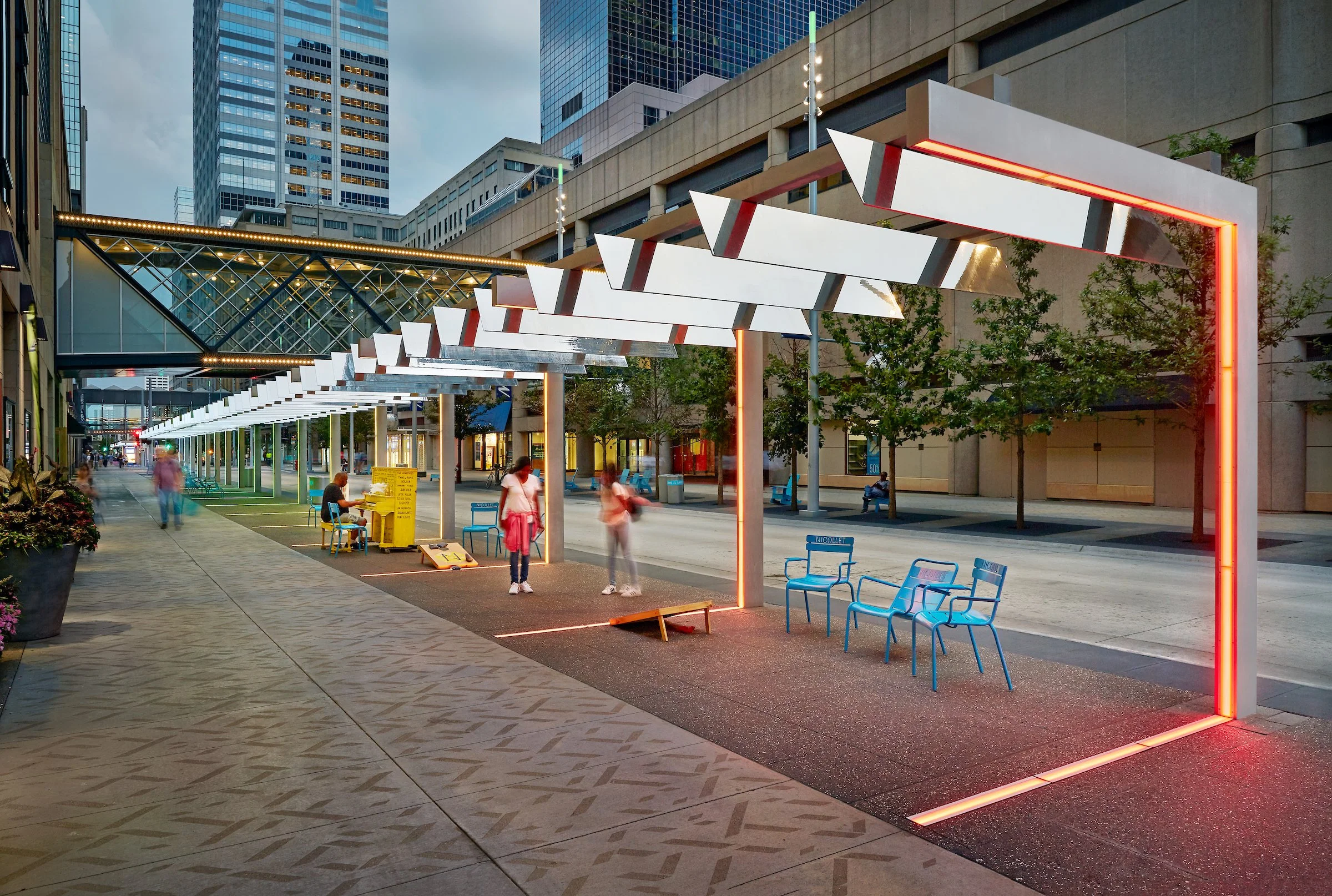 Urban street scene with modern architecture, illuminated display structures, and people walking and sitting on blue chairs during dusk.