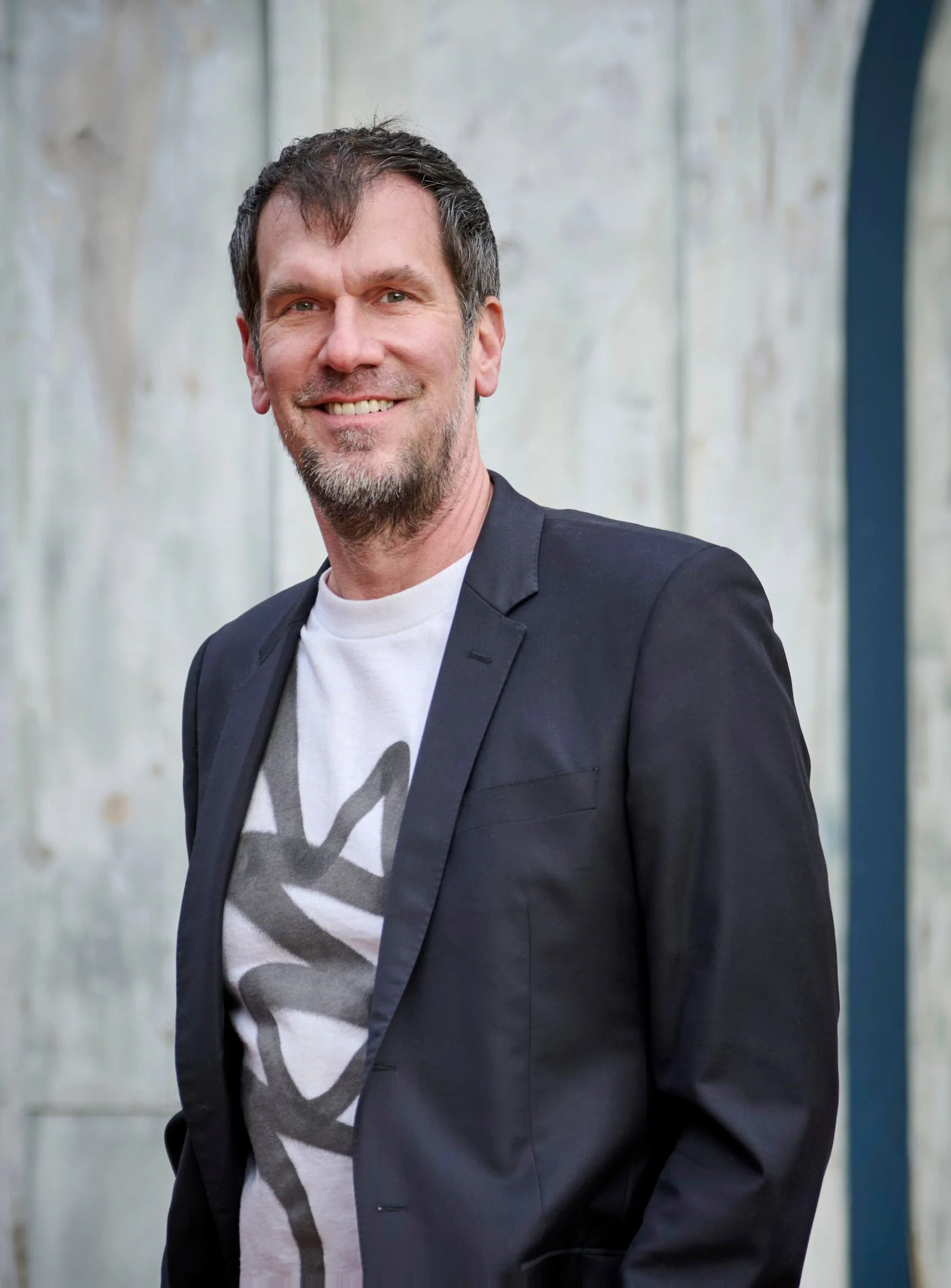 Portrait of a man—waist up—wearing a white graphic tee under a navy blazer, hands in pockets, in smiling at camera.