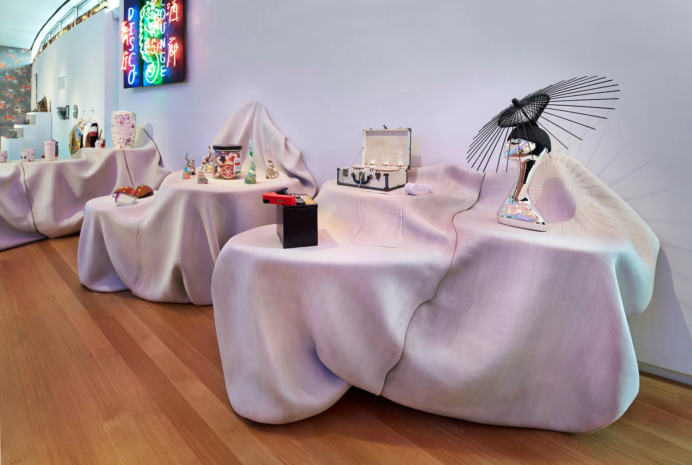 Display of eclectic decorative objects arranged on draped fabric-covered tables, including a black parasol, ceramic sculptures, a jewelry box, and neon sign art, inside a modern gallery with wooden flooring.