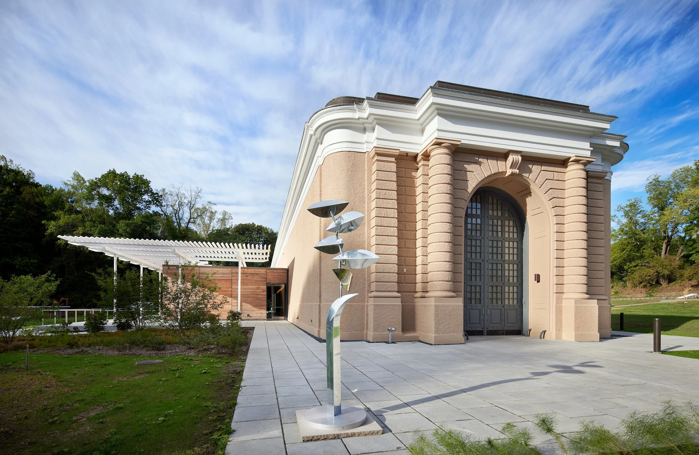 A historic building with large arched door and detailed stonework, adjacent to modern outdoor sculpture, surrounded by green landscape and a partly cloudy sky.