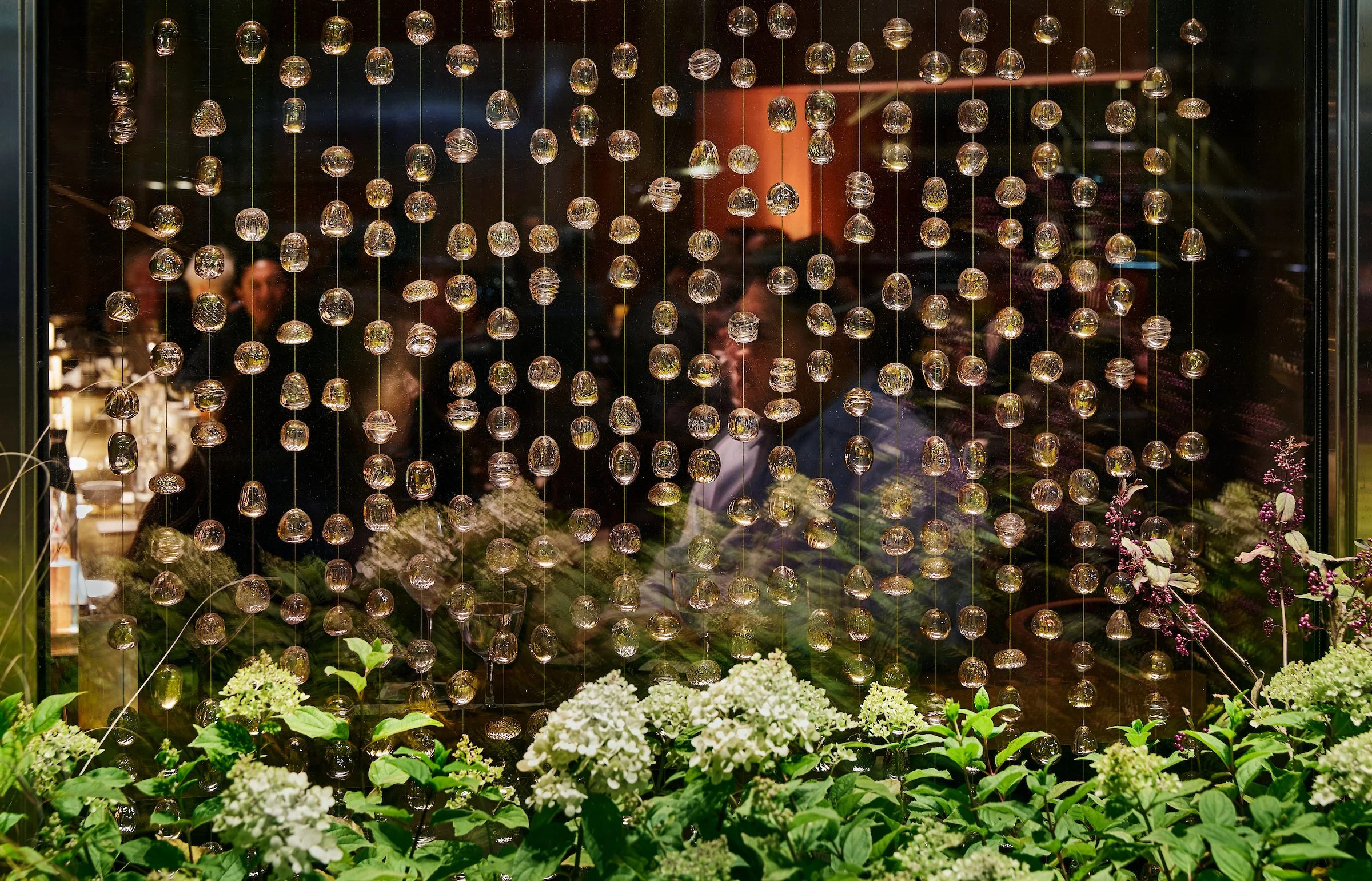 Decorative glass window with hanging glass orbs, floral arrangements and people reflected in the background.