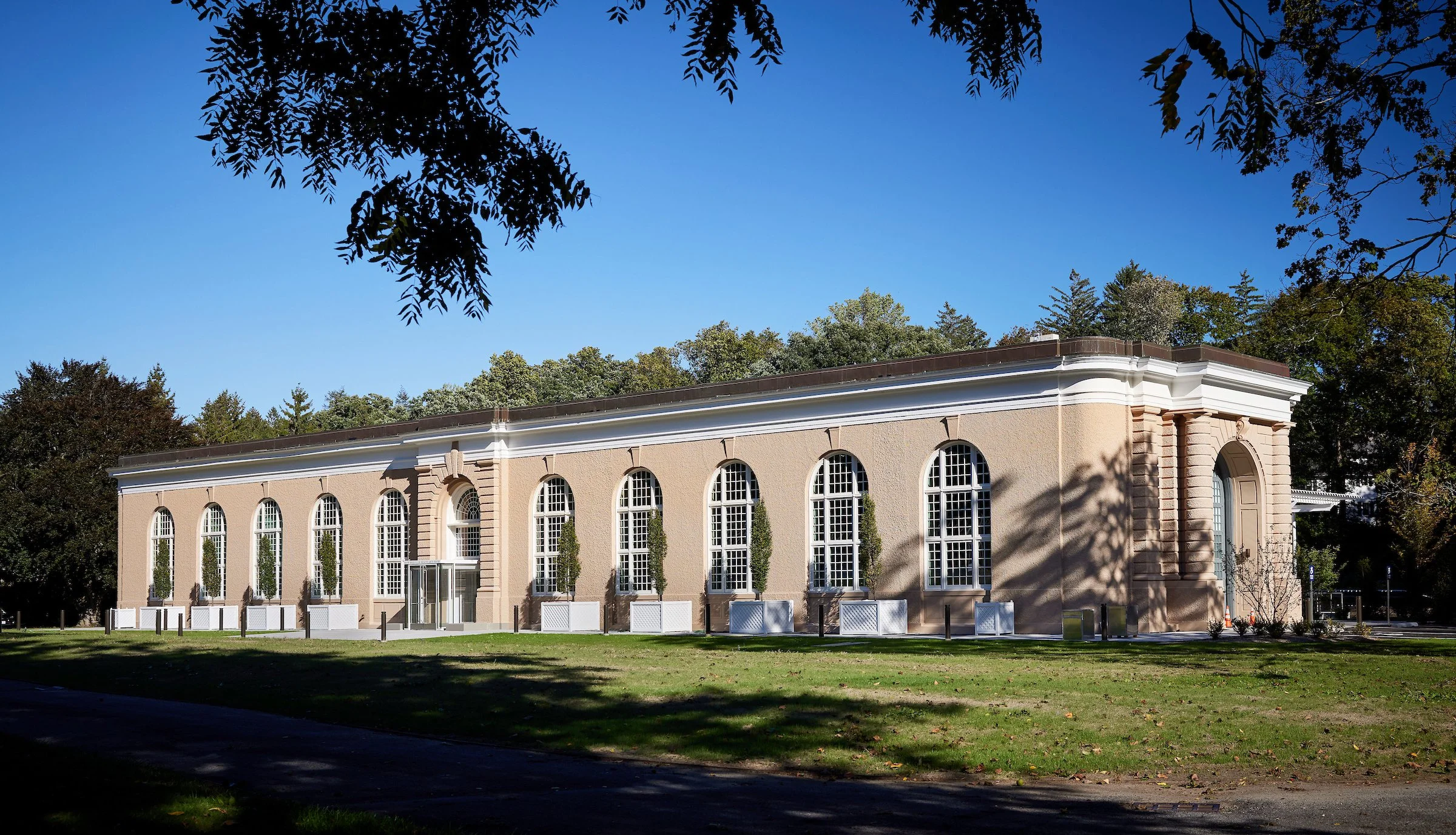 A large, historic beige building with tall arched windows and decorative architectural details, set in a park-like area with grass and trees.
