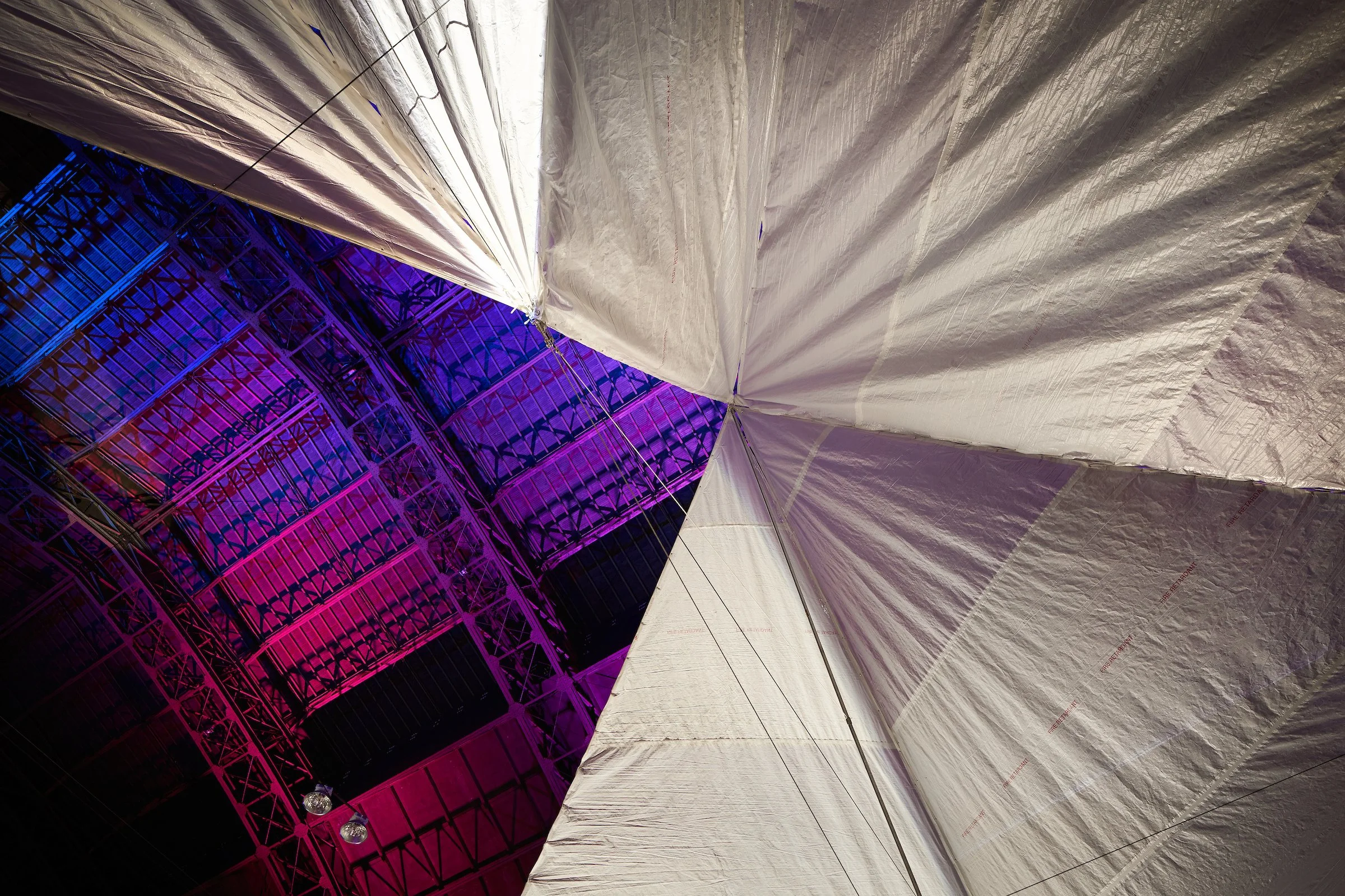 Looking up at the inside of a large industrial or event tent with a white fabric ceiling and purple lighting on a building structure visible through the opening.