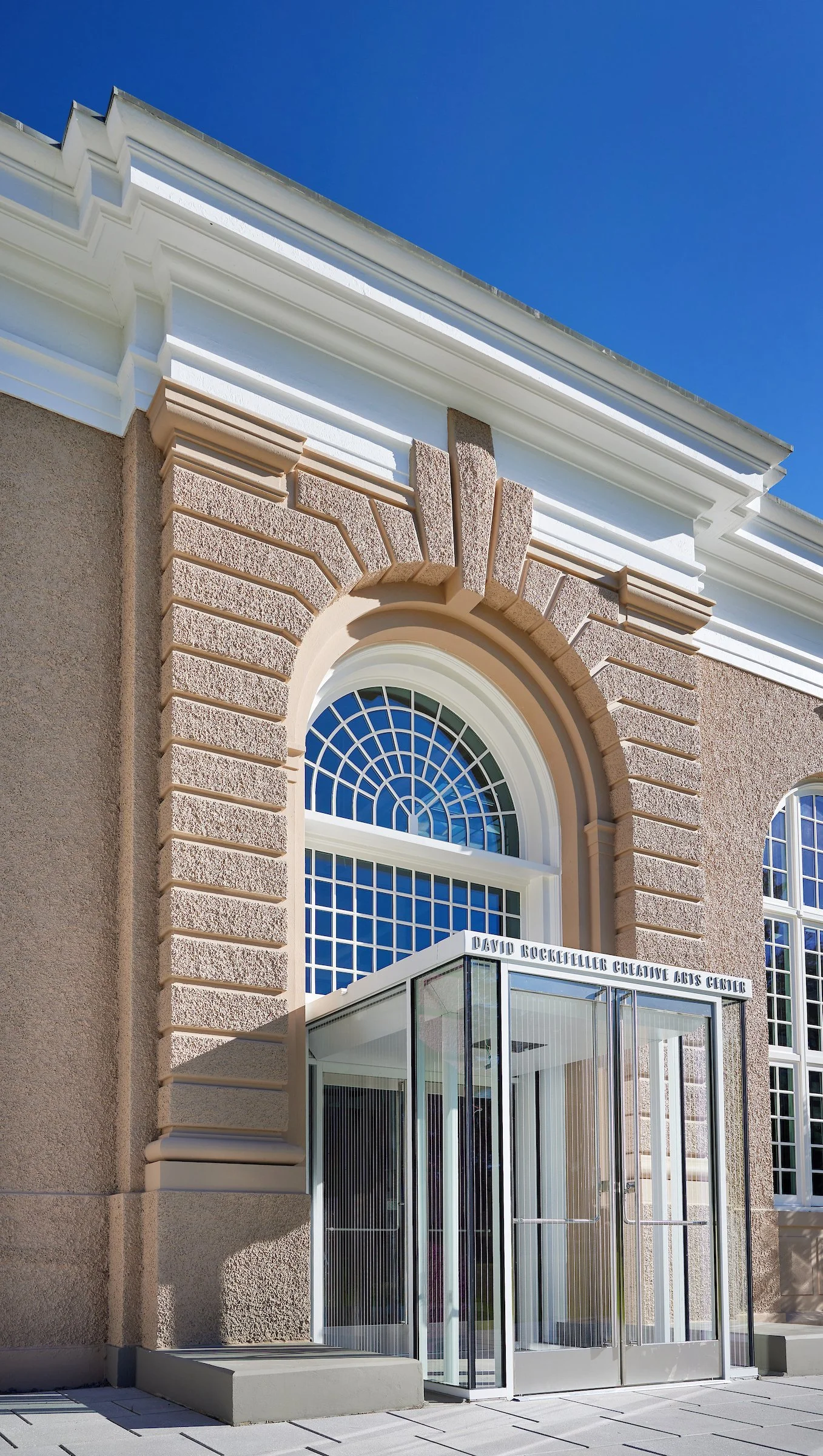 Entrance of the David Rockefeller Creative Arts Center with glass doors and arched window, brick and stucco facade, clear blue sky.