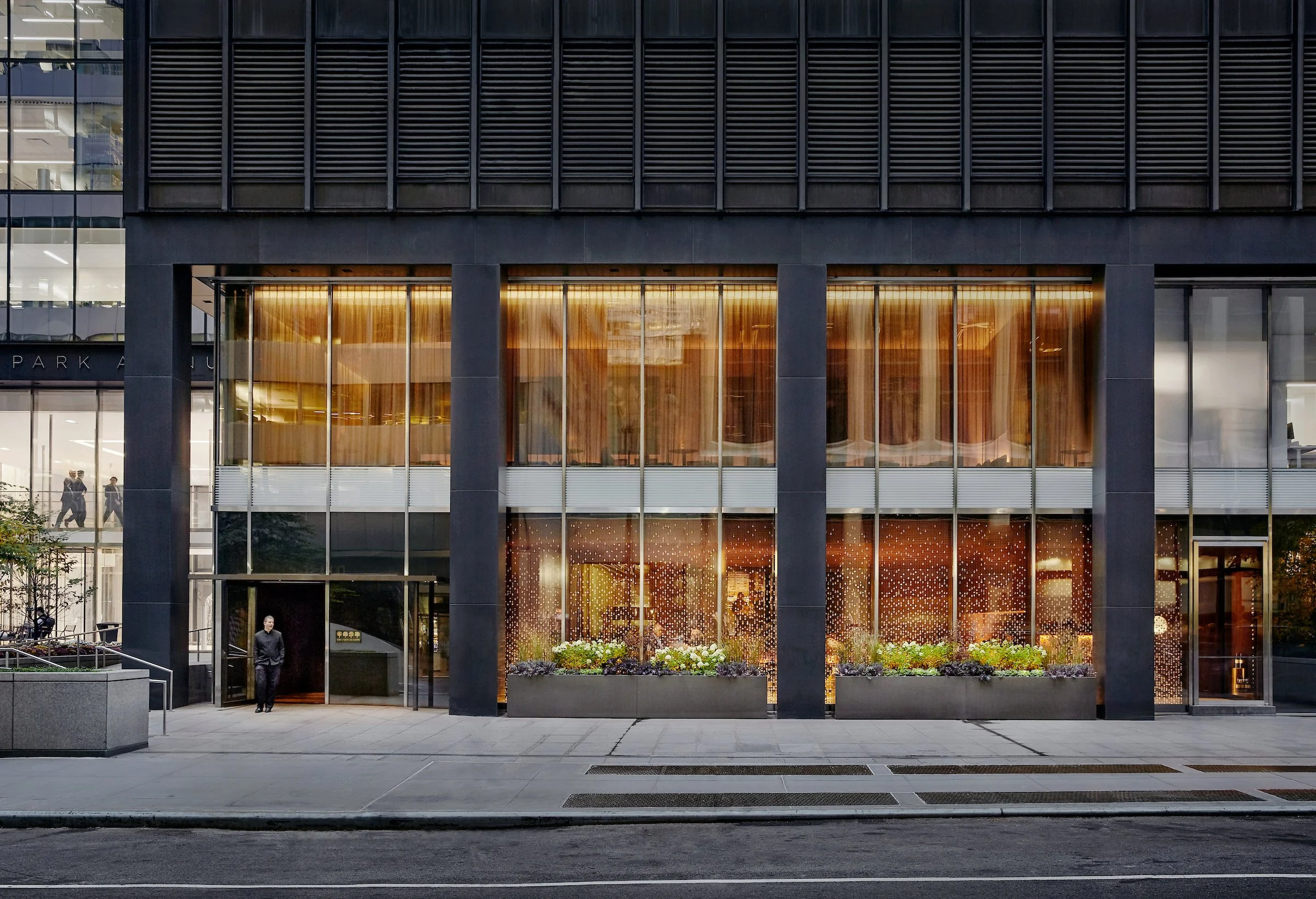 Modern multi-story office building with large glass windows and black exterior panels. The ground floor features decorative lighting inside and flower planters outside the entrance. A man in black clothing is walking out of the entry, with a sidewalk