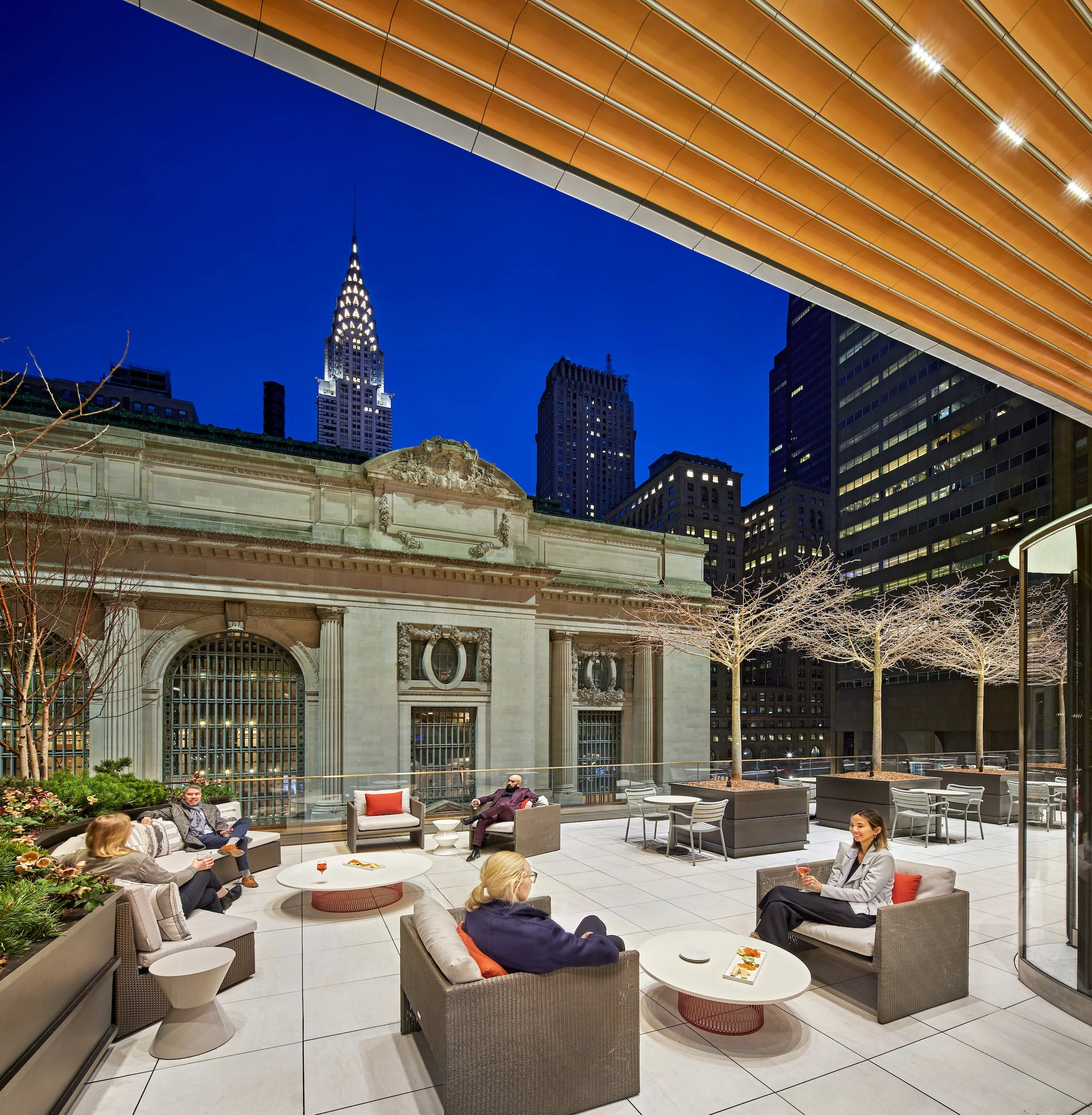 Outdoor rooftop lounge at night with people sitting on sofas, drinking wine, and talking, with city buildings and the Empire State Building in the background.