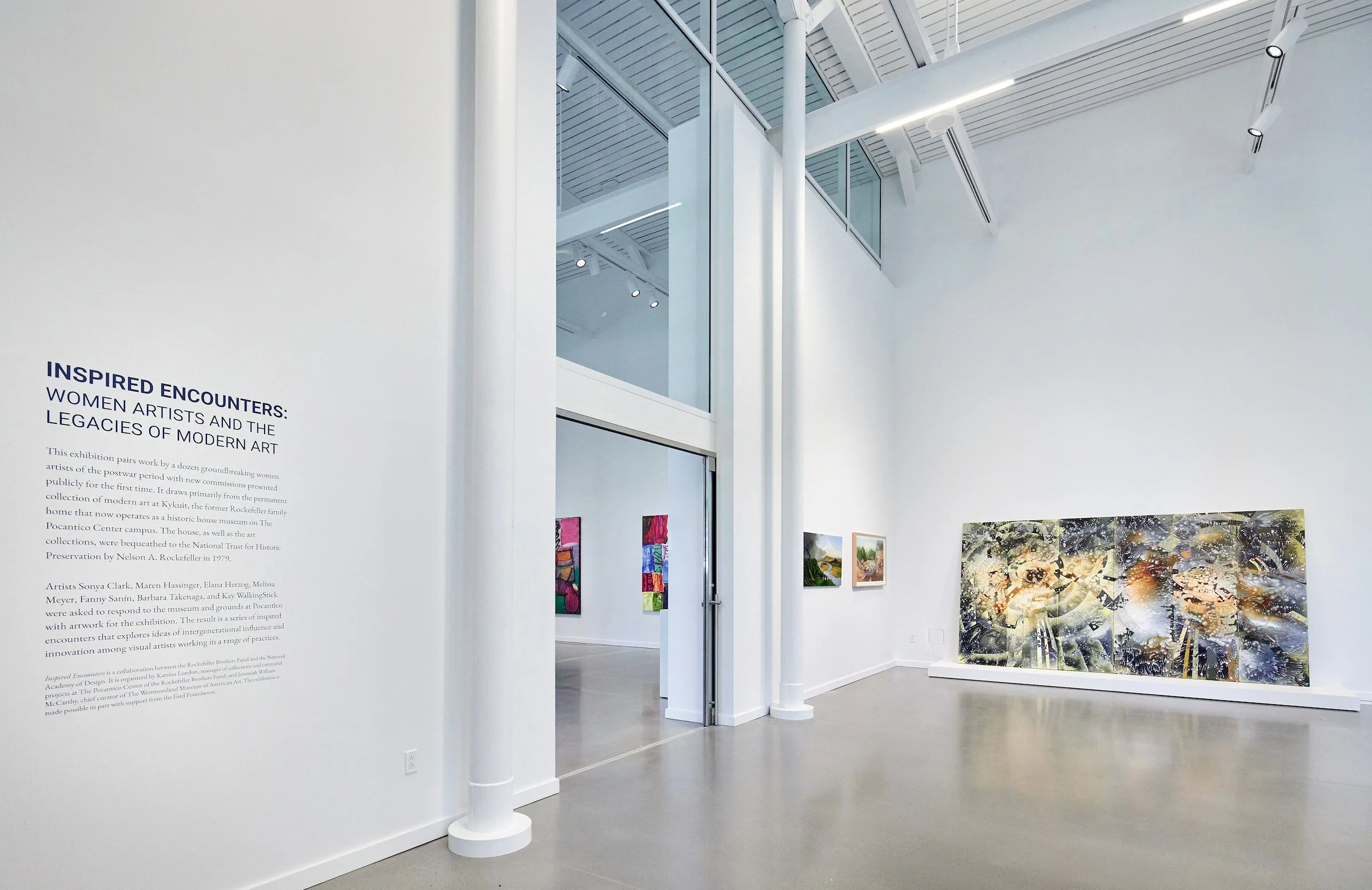 Art gallery with white walls and high ceilings, displaying colorful modern paintings; text panel on left side explaining the exhibit.