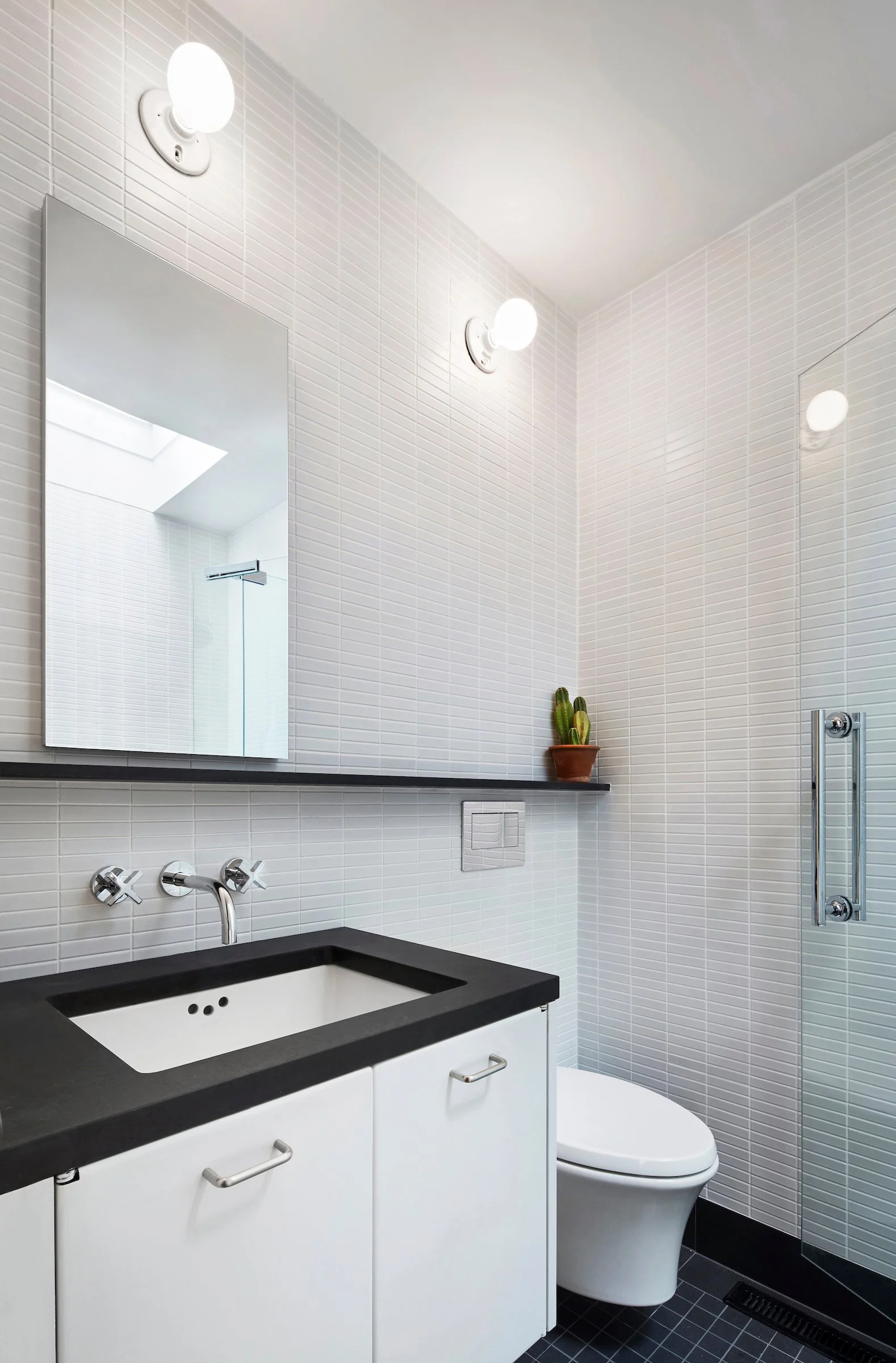 Modern minimalist bathroom with white tiled walls, black countertop sink, white cabinet, toilet, mirror, two round wall lights, small potted cactus on black shelf, and glass shower partition.