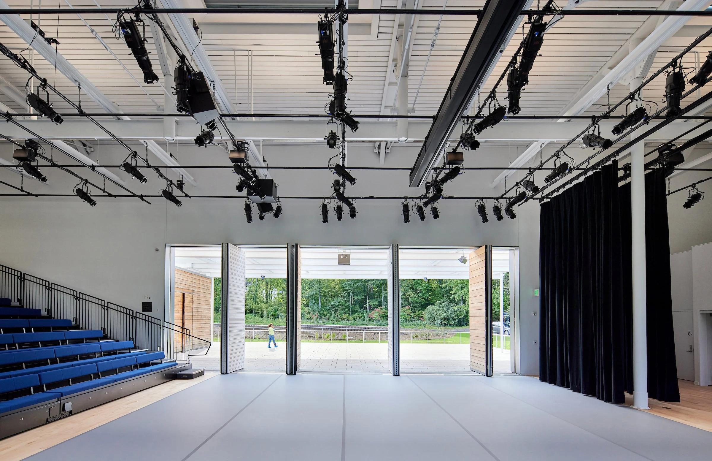 Empty stage with black curtains, blue seating, and stage lights, with open doors leading outside to a grassy area with trees and a person walking.
