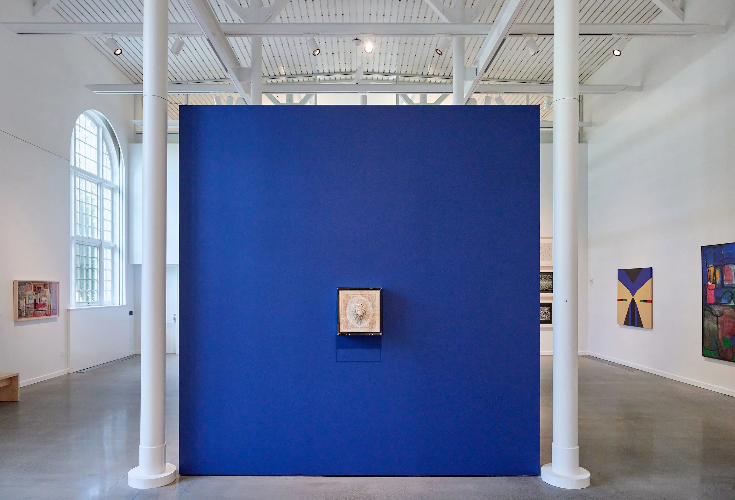 An art gallery with white walls and a blue partition wall in the center. Four paintings are visible on the walls, with one framed artwork on the blue wall.