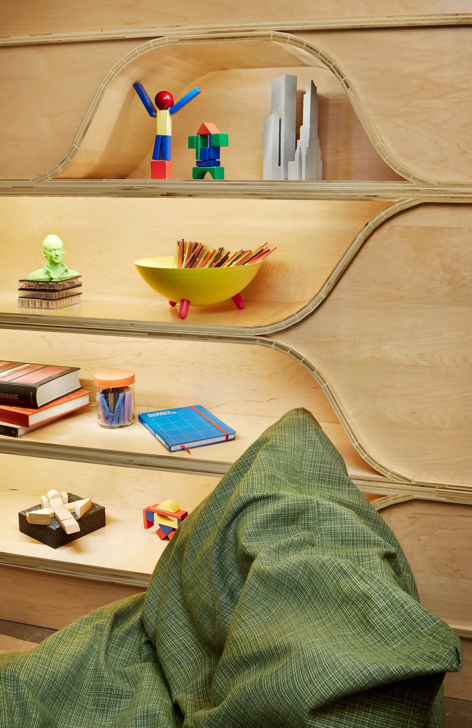 View of a wooden shelf with various colorful toys, books, and decorative objects, and a person's green pants in the foreground.