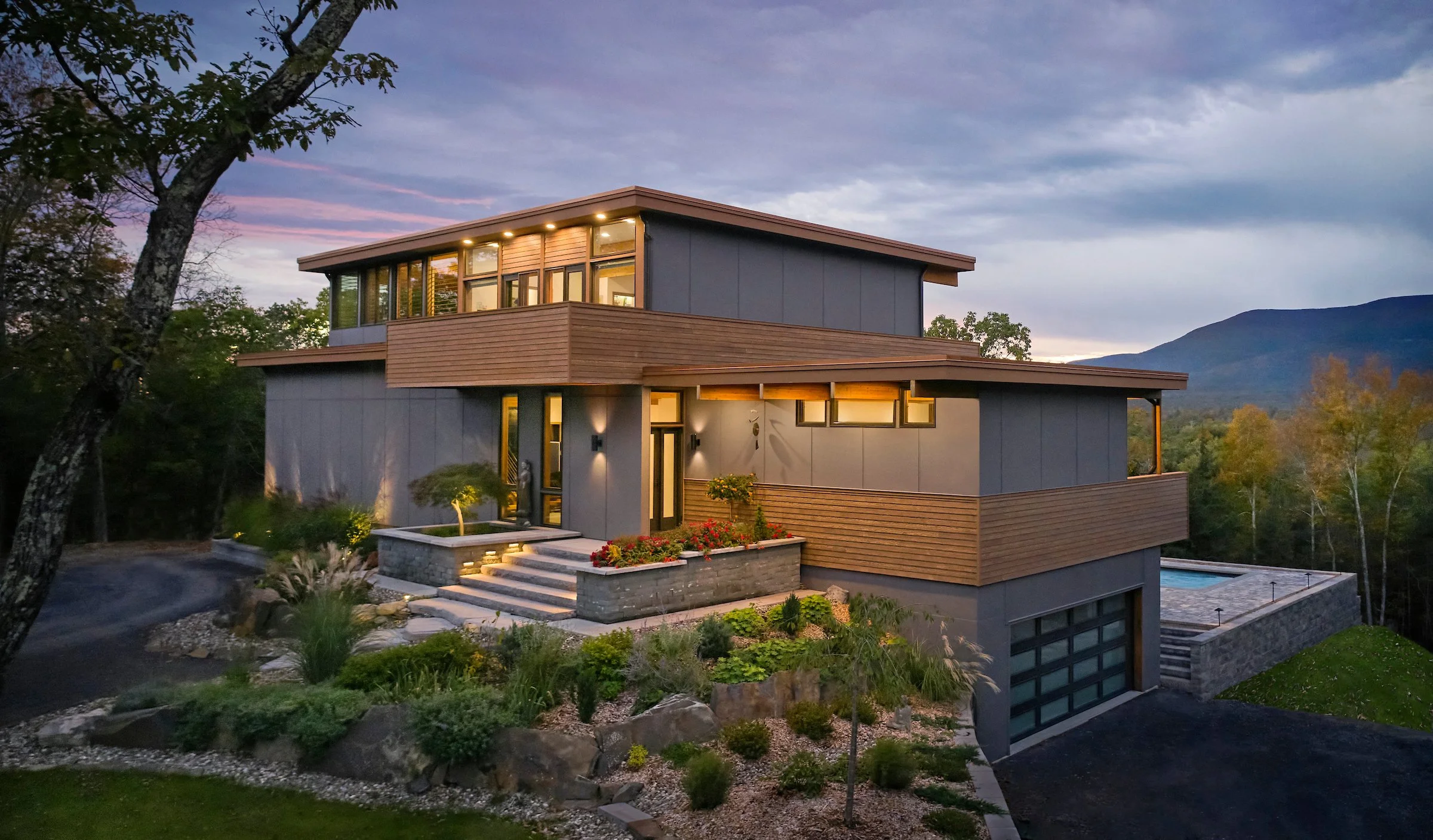 Modern multi-level house with large windows, wooden accents, landscaped front yard, and mountain view in the background during dusk.