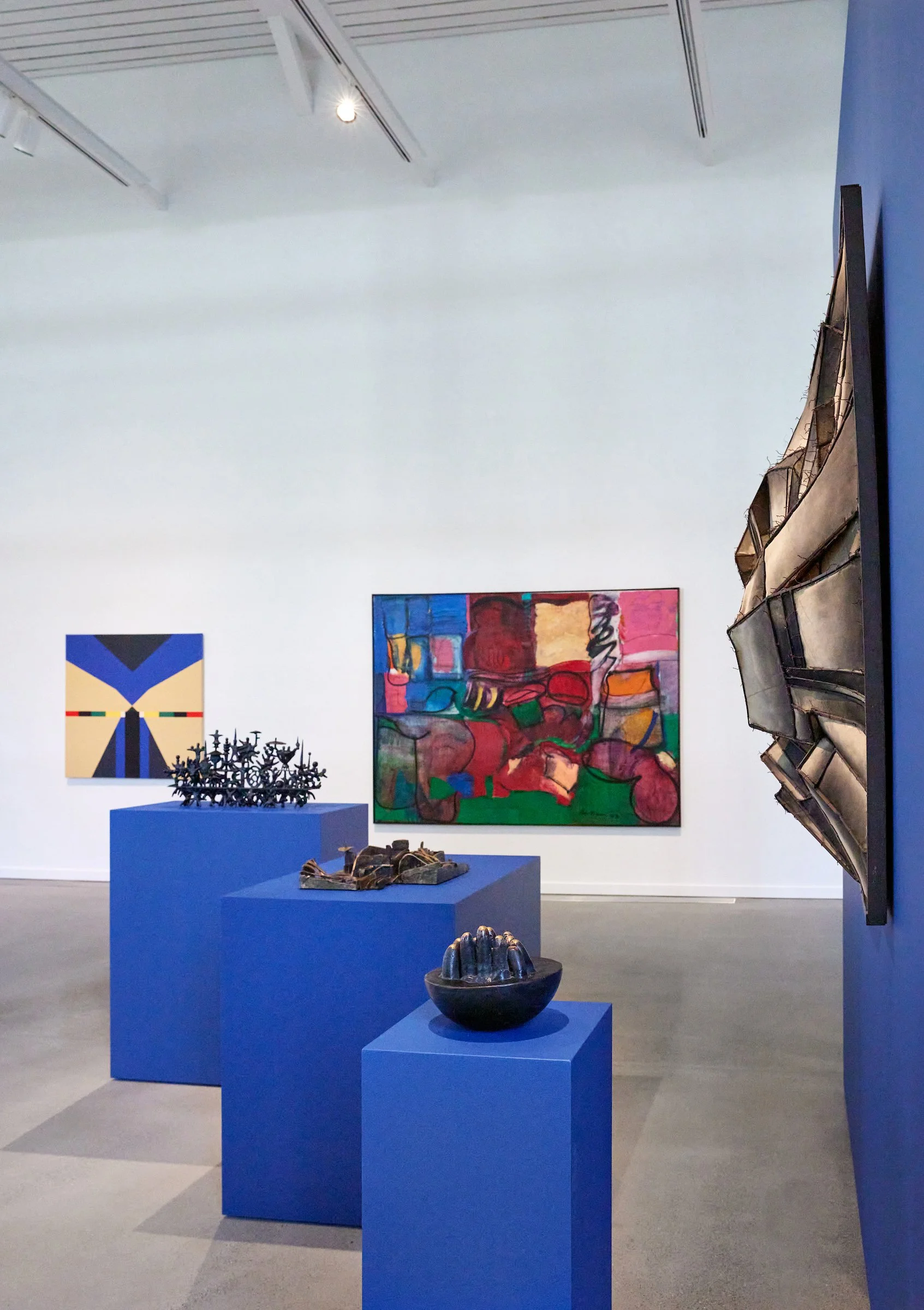 Art gallery with abstract paintings on the white wall, black sculptures on blue pedestals, and a crumpled metallic sculpture on a pedestal, with modern ceiling track lighting.