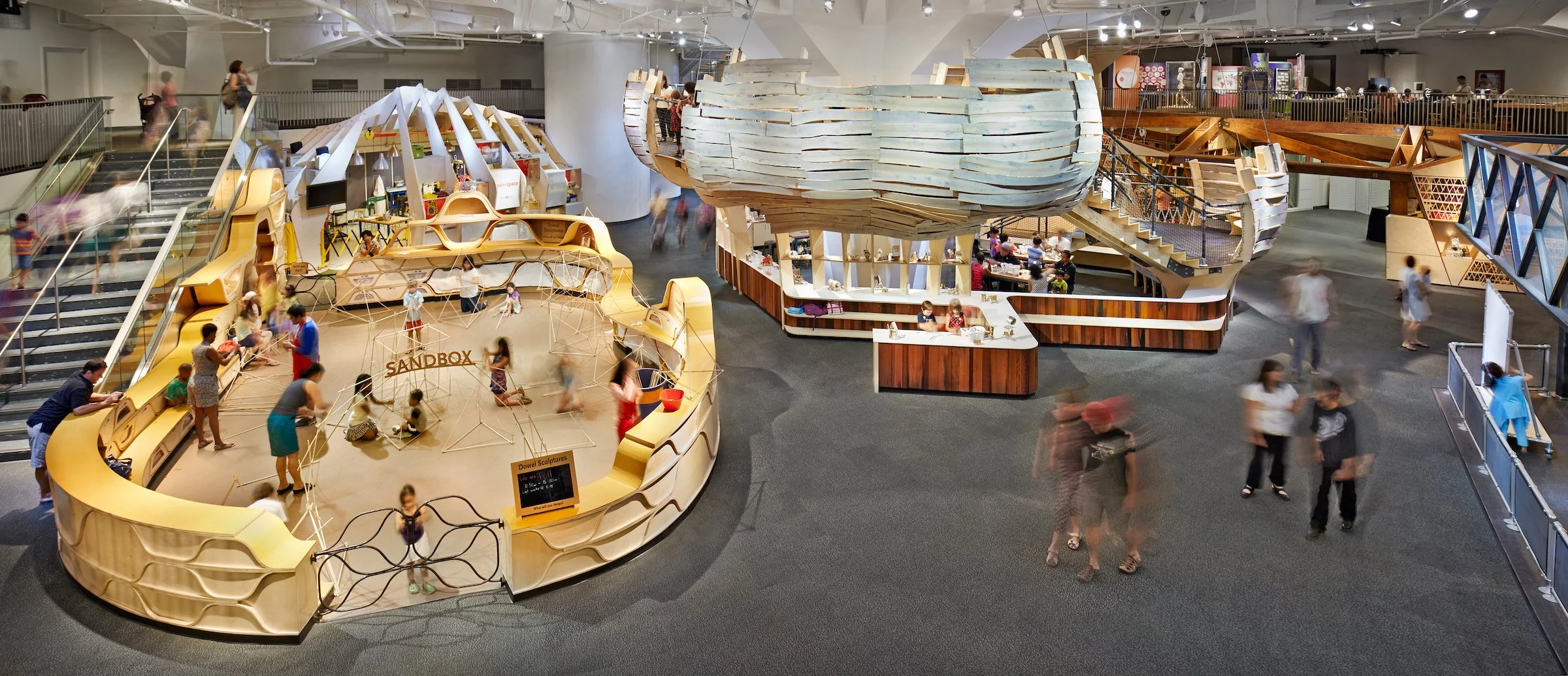 Indoor children's play area with a sandbox labeled 'SANDBOX', climbing structures, and multiple levels including a wooden balcony and stairs, with children and adults walking around and engaging in activities.