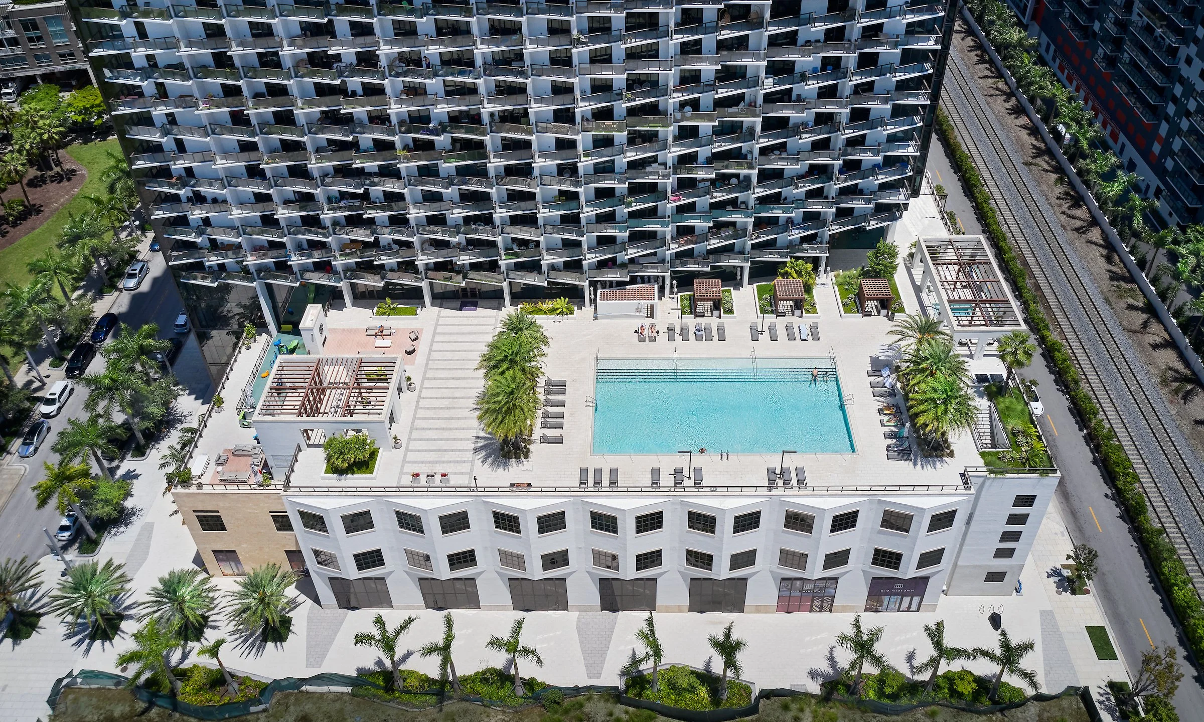 Aerial view of a modern apartment complex with a rooftop swimming pool, surrounded by palm trees and outdoor lounging areas.