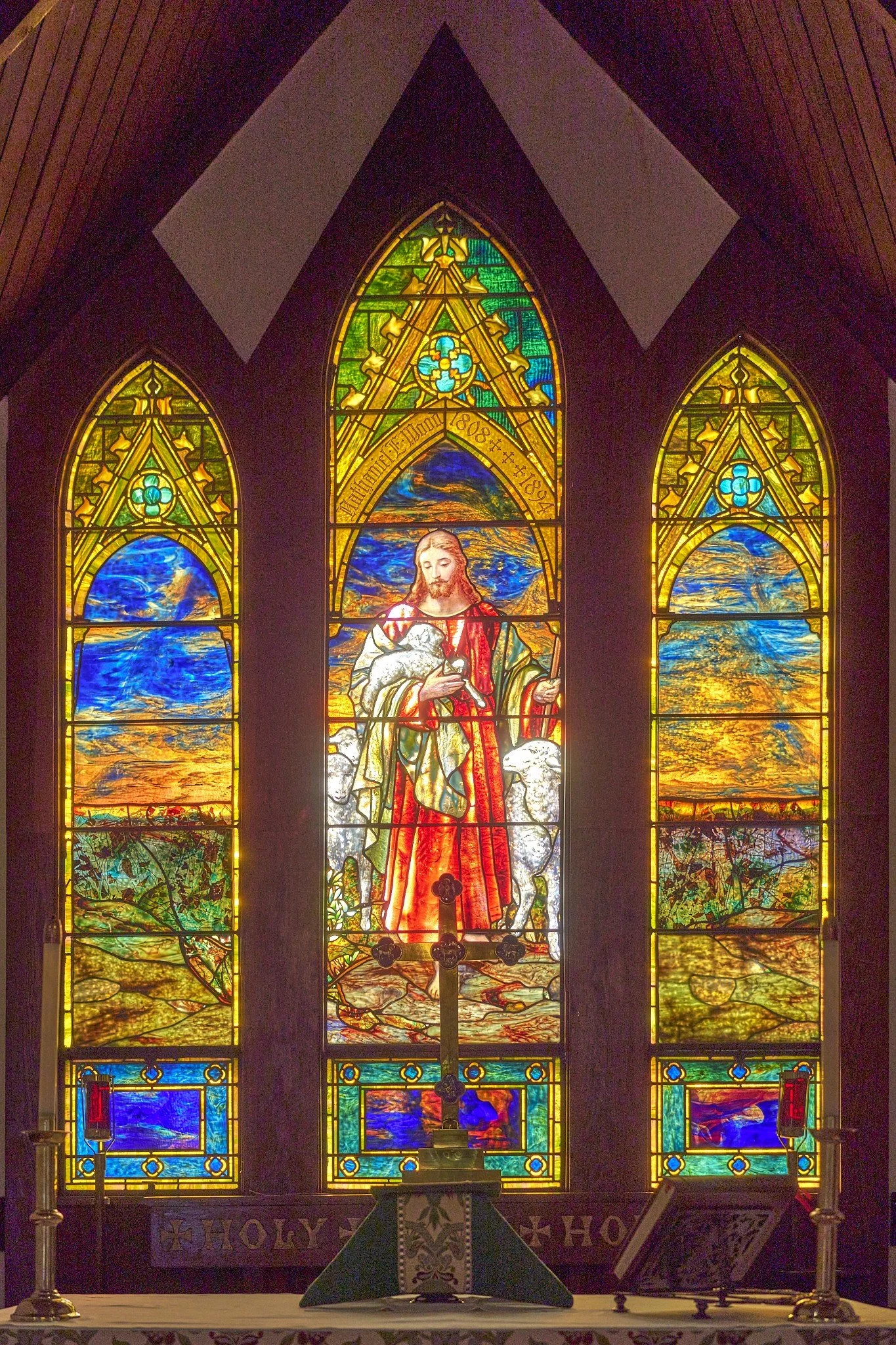 Colorful stained glass window depicting Jesus Christ holding a lamb, surrounded by scenic landscape scenes and a rainbow, in a church setting with an altar and candles.