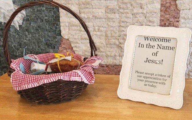 An Easter basket with a red and white checkered cloth, containing small wrapped gifts, and a framed sign that reads, "Welcome In the Name of Jesus! Please accept a token of our appreciation for your presence with us today," on a wooden table against a brick wall.