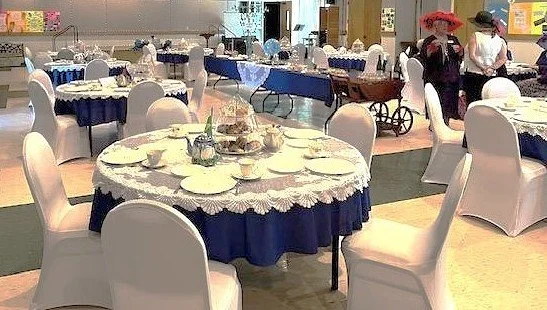 A banquet hall with multiple round tables covered with blue tablecloths and white chair covers, set for a meal with plates, cutlery, and centerpieces.