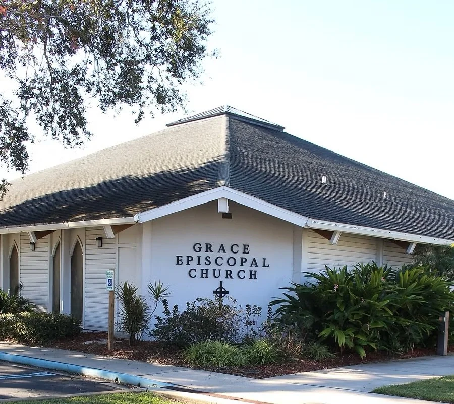 Front view of Grace Episcopal Church with a white exterior, dark roof, and landscaping in the front, including bushes and small trees.