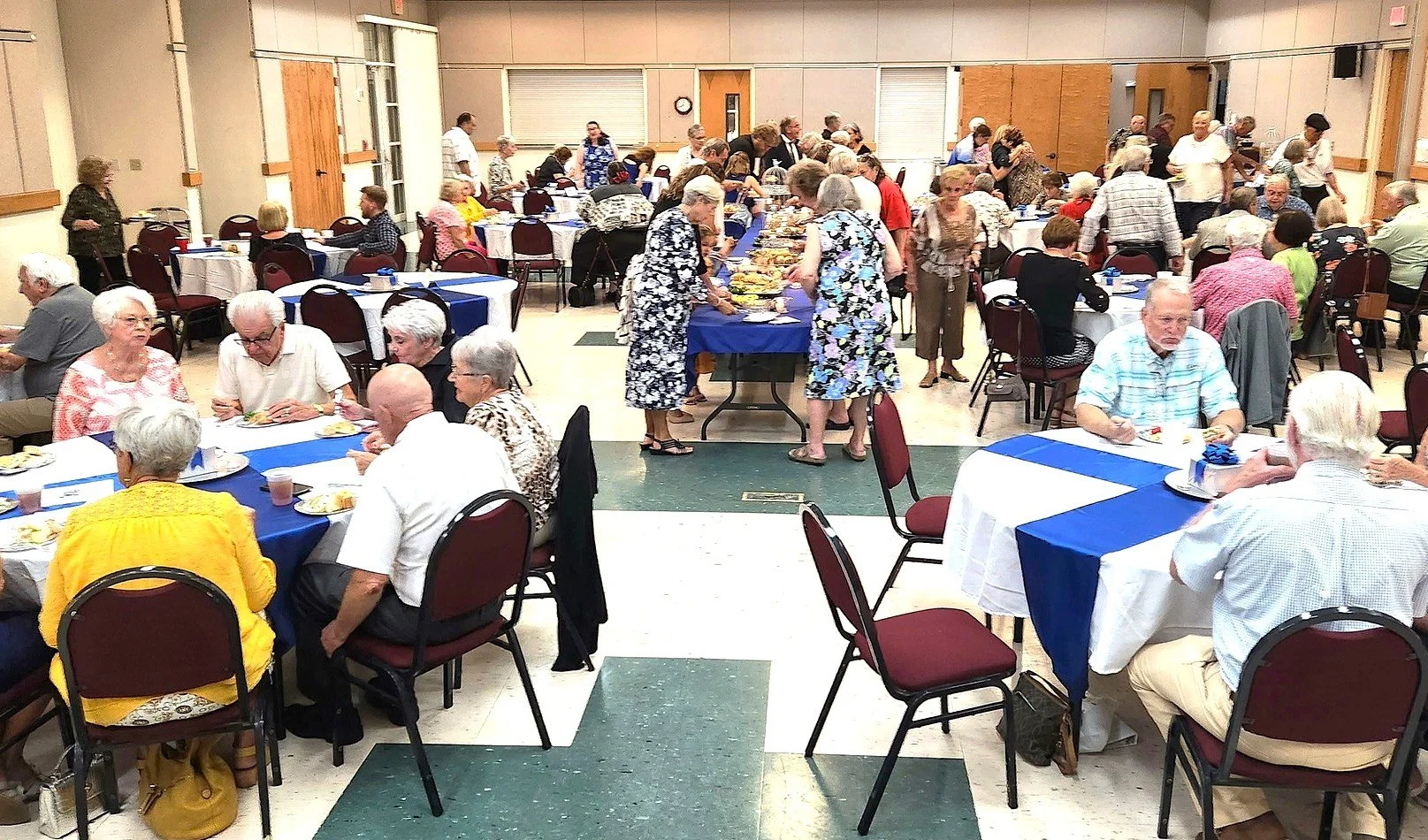 A large gathering of people at a community event in a hall. Some are sitting at round tables covered with blue and white tablecloths, eating and talking. Others are standing around a buffet table with food. The hall has beige walls, wooden doors, and a green and white floor.