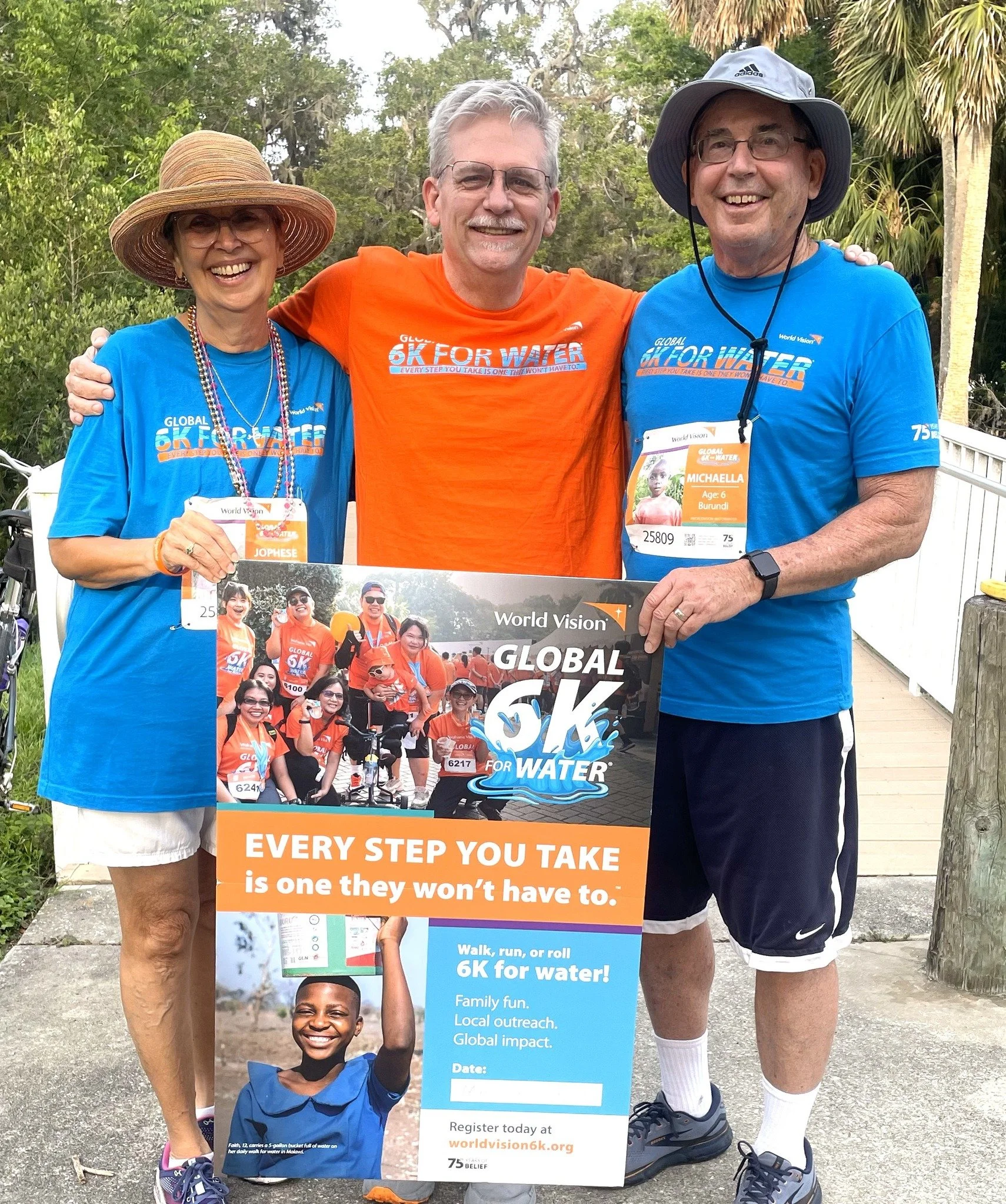 Three people standing outdoors, smiling and holding a large sign promoting a water charity event. They wear bright blue and orange shirts with event logos, and two of them have race bibs. The background includes trees and a wooden walkway.