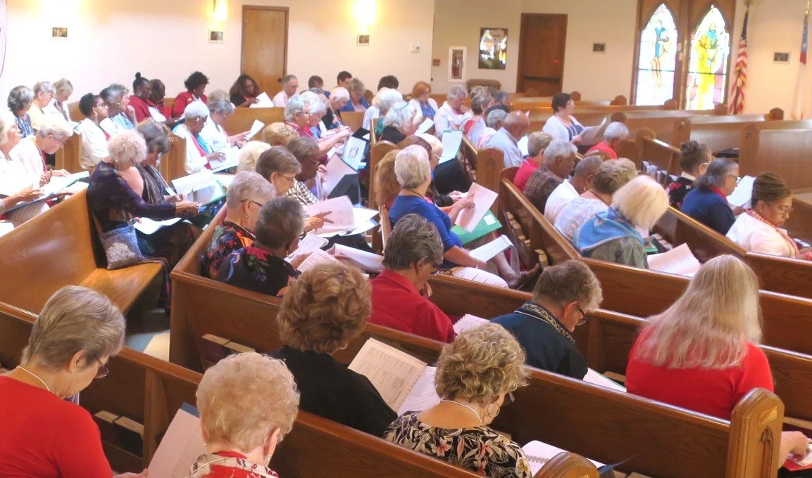 A congregation of people seated in wooden pews inside a church, reading and singing from hymn books or programs.