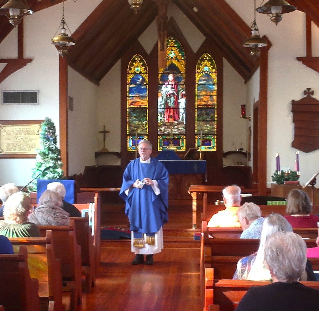 Reverend leading a worship service in a small church with stained glass windows behind him, an altar, and pews filled with congregants.