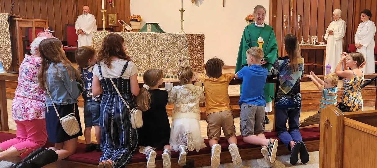 Children kneeling at the front of a church during a religious service, with a priest and altar in the background.