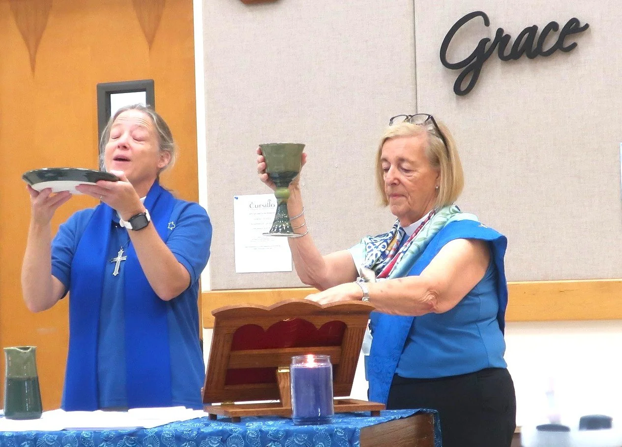 Two women standing at a table, engaged in a religious or spiritual activity. One woman holds a plate, and the other holds a chalice, with a candle and a wooden box on the table.