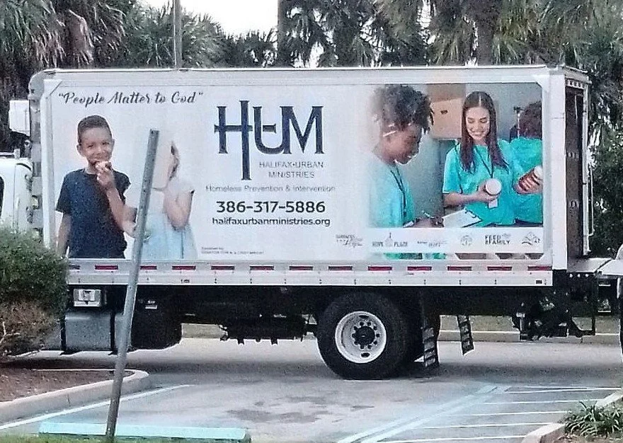 A truck with outdoor advertising for Halifax Urban Ministries. The ad features children, one boy smiling and two girls in blue shirts, engaging in ashy activities. The text includes the phrase "People Matter to God" and the organization’s contact information.