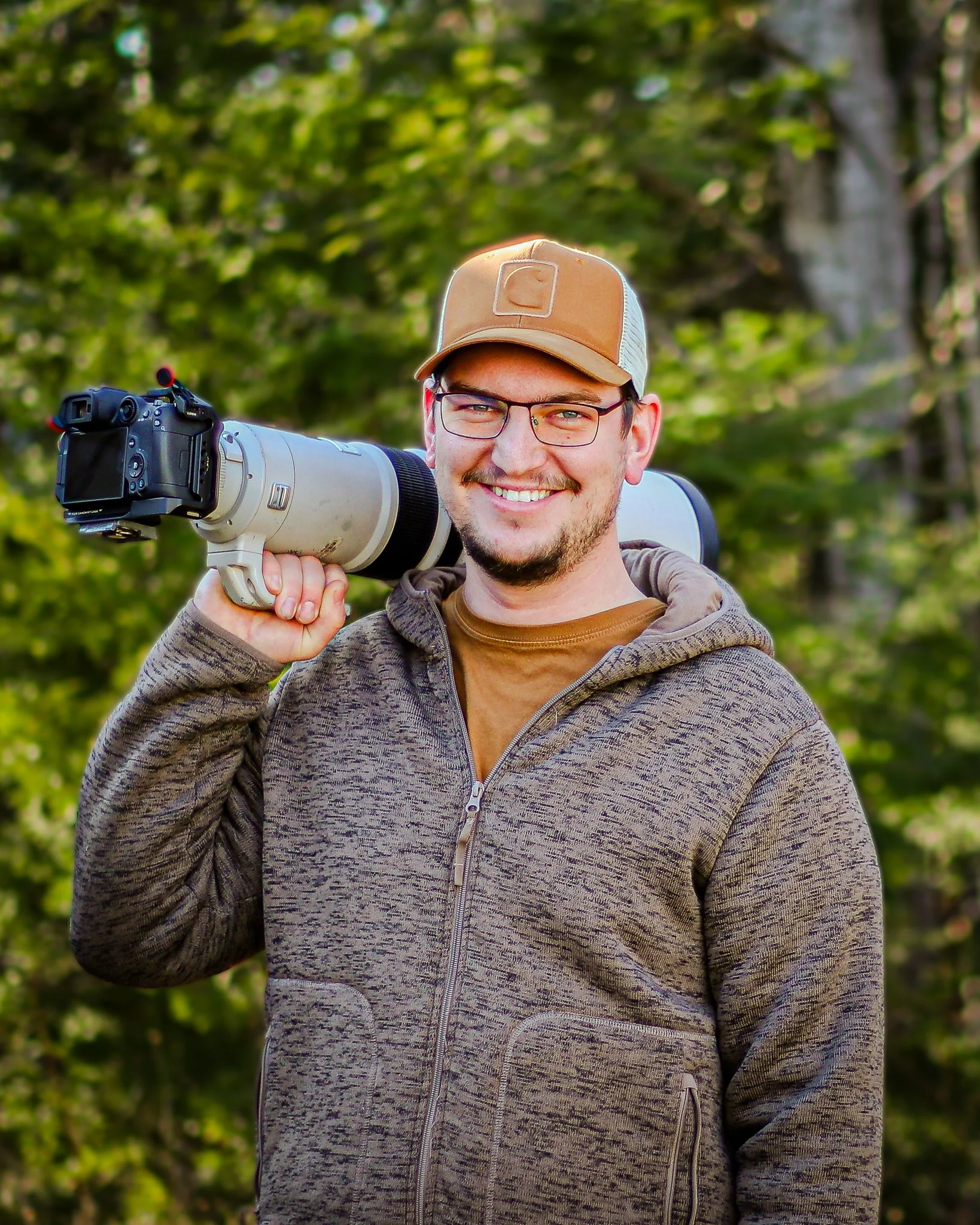 A smiling man wearing glasses and a cap, carrying a large camera with a telephoto lens on his shoulder in a forest setting.