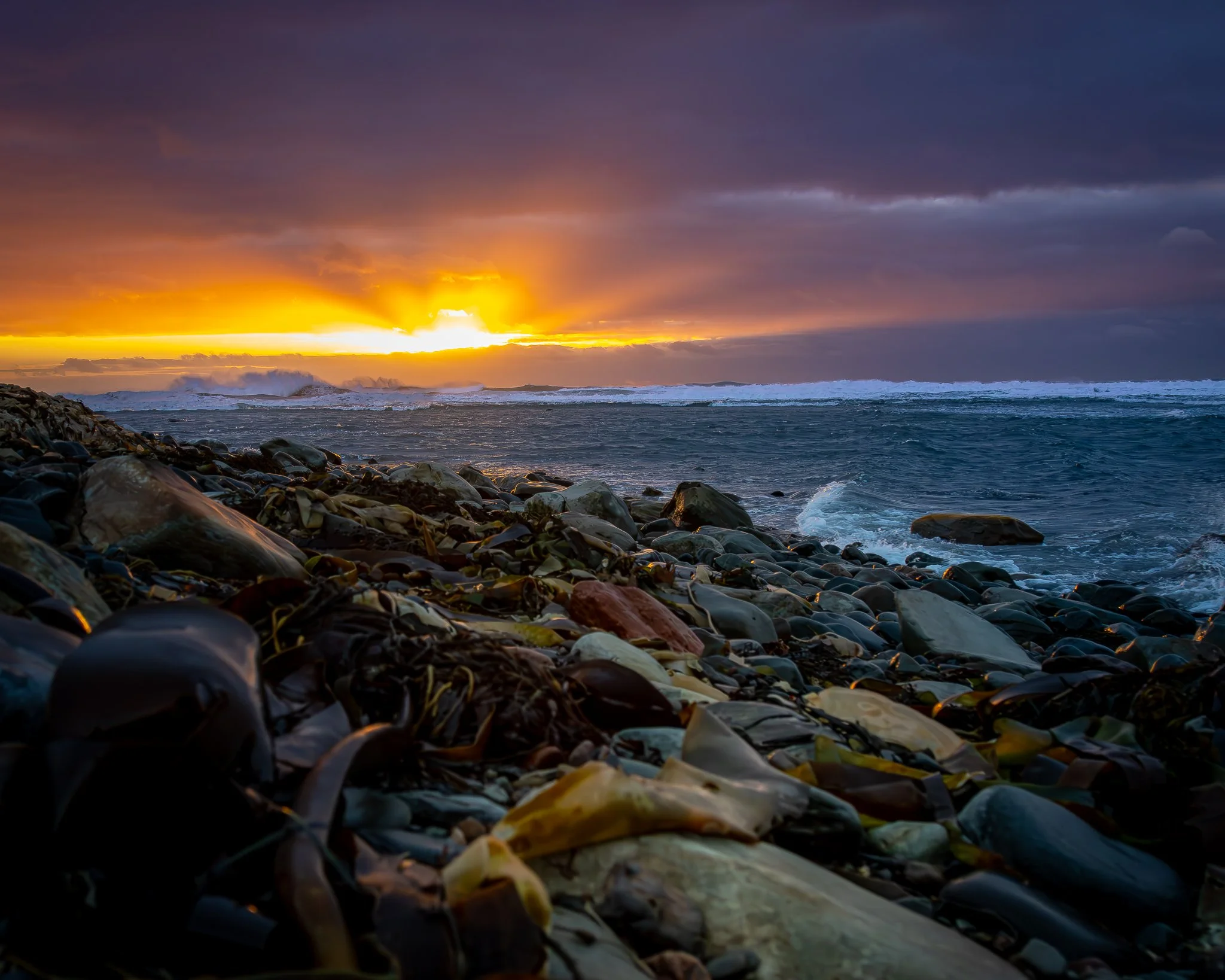 Sunset over a rocky shoreline with waves crashing and dark clouds in the sky.
