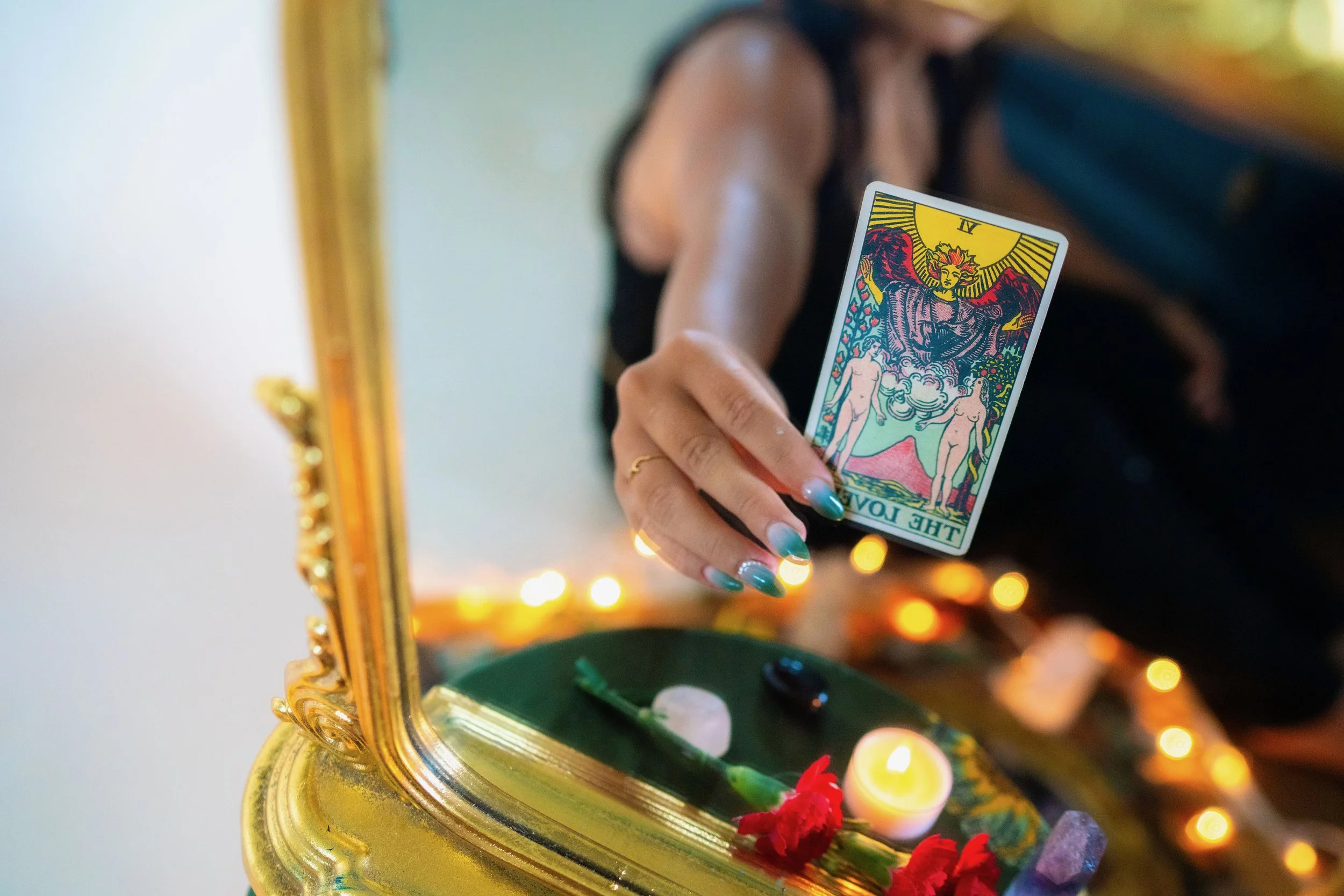 A person holding a tarot card labeled 'The Fool' in front of a mirror with gold frame decoration. There is a small candle, flowers, and stones on a green surface in front of the mirror, and the person appears to be performing a tarot reading or ritual.