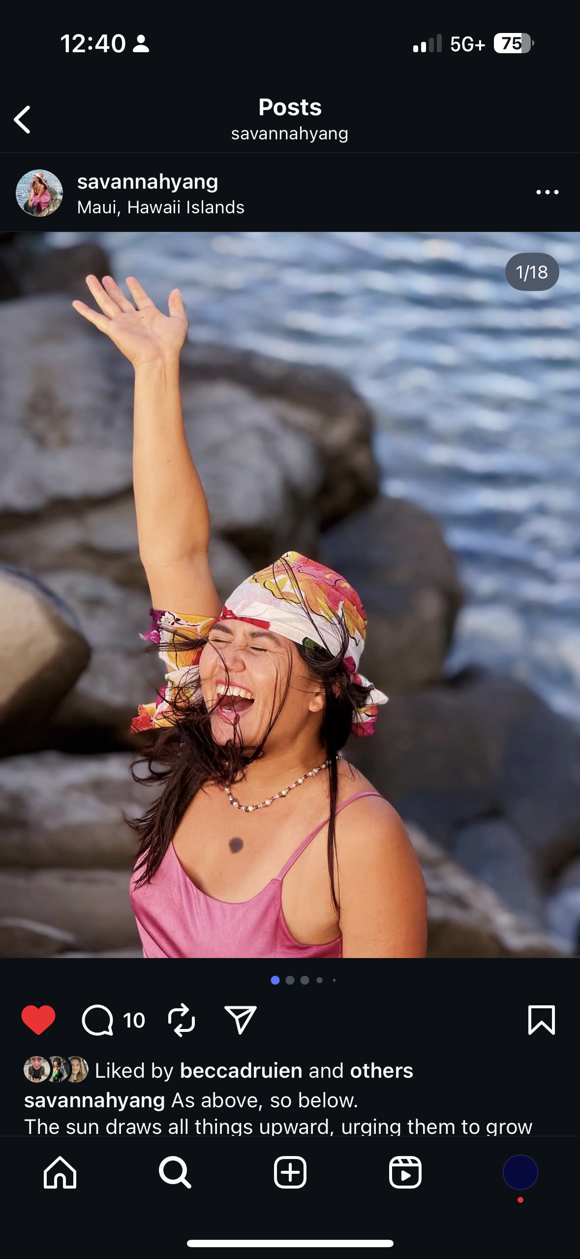 A woman wearing a colorful headscarf and pink dress, smiling and raising her arm joyfully near rocks and water on a beach in Maui, Hawaii.