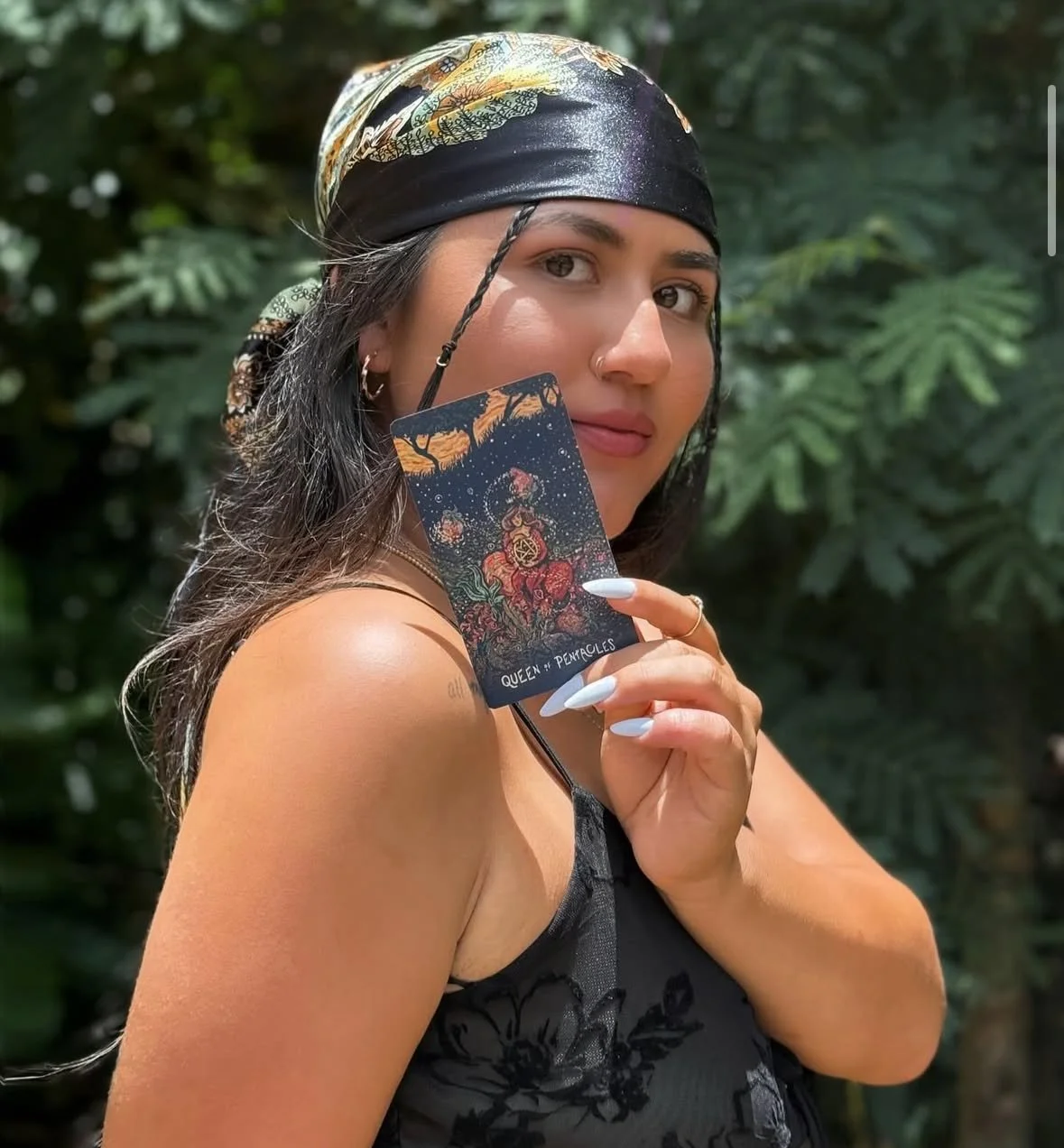 A woman with long dark hair, wearing a black bandana and black top, holds a tarot card labeled 'Queen of Pentacles' in front of a green leafy background.