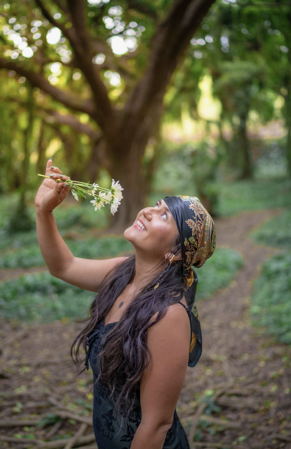 A woman with long hair wearing a black sleeveless dress and a colorful headscarf holding a small bouquet of white flowers, smiling and looking up with a large tree and greenery in the background.