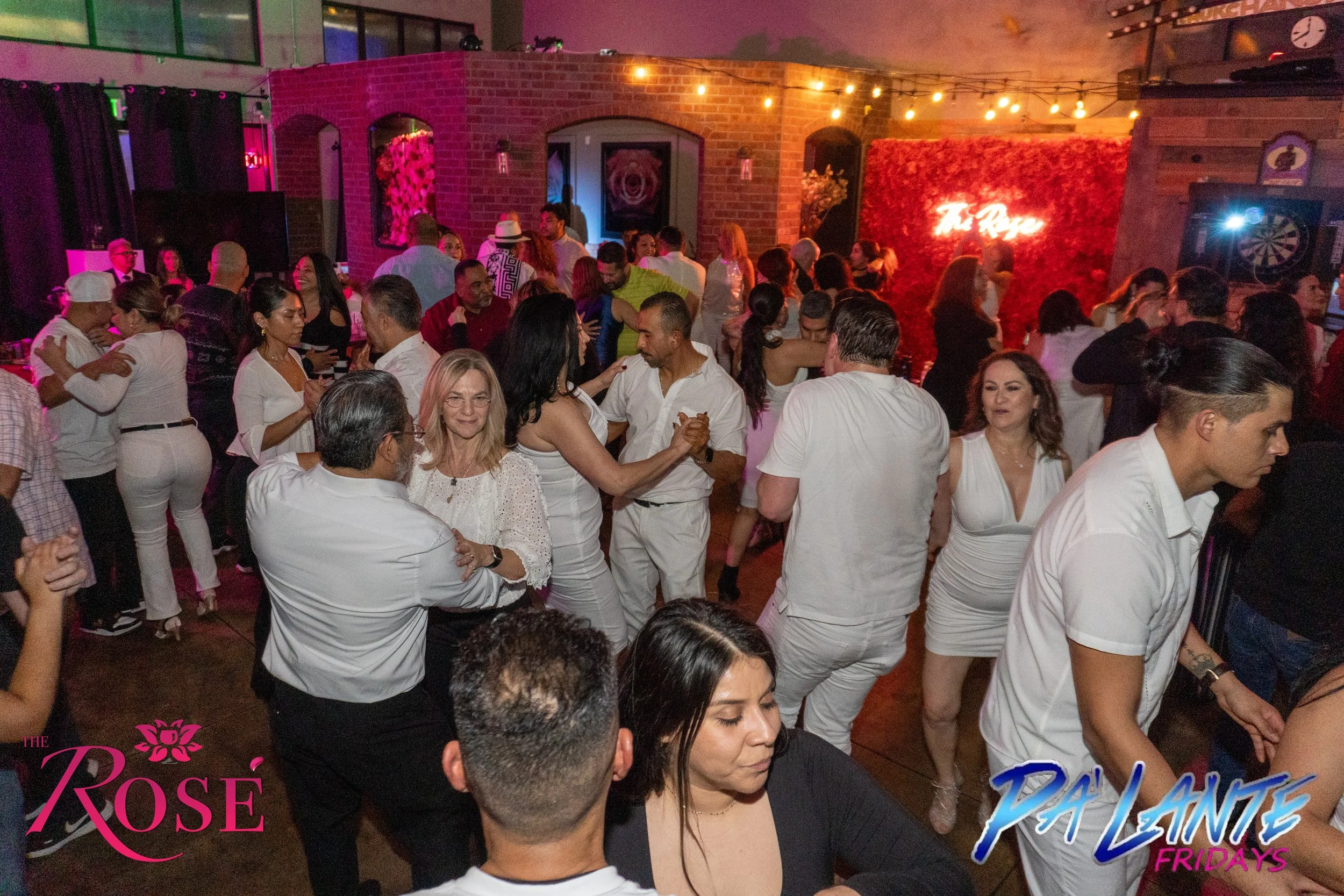 People Salsa dancing and socializing at a nightclub with brick walls, string lights, and a red floral backdrop with neon sign reading 'Tequila.'