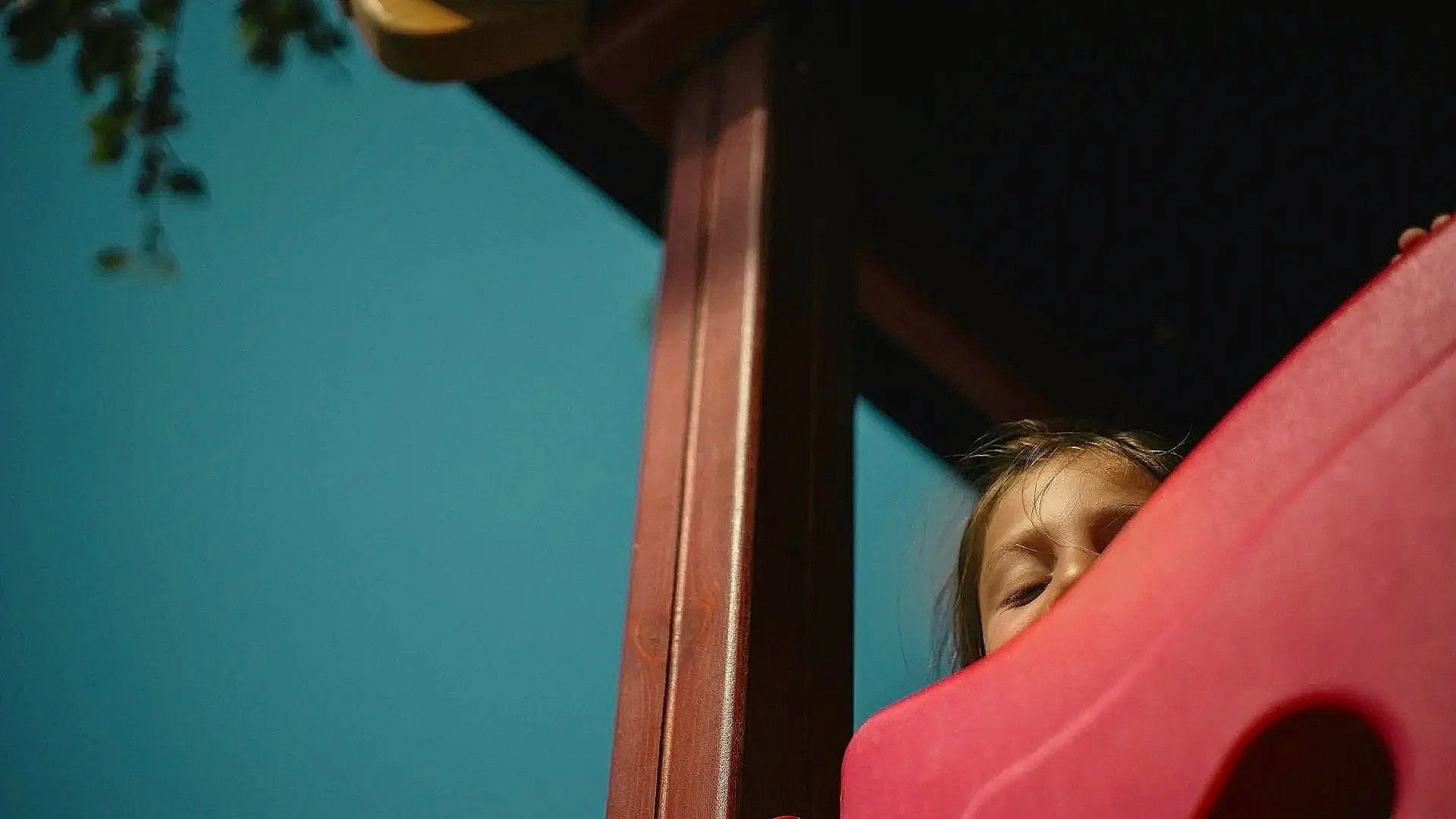 Child peeking behind a pink object, with half of their face and eye visible, indoors with a wooden structure and a blue wall in the background.