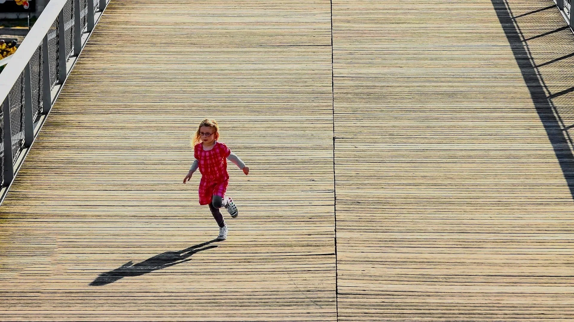 A young girl with glasses running on a wooden pier with a white railing on the left side, casting a shadow. She is wearing a pink checkered dress, glasses, and gray pants.