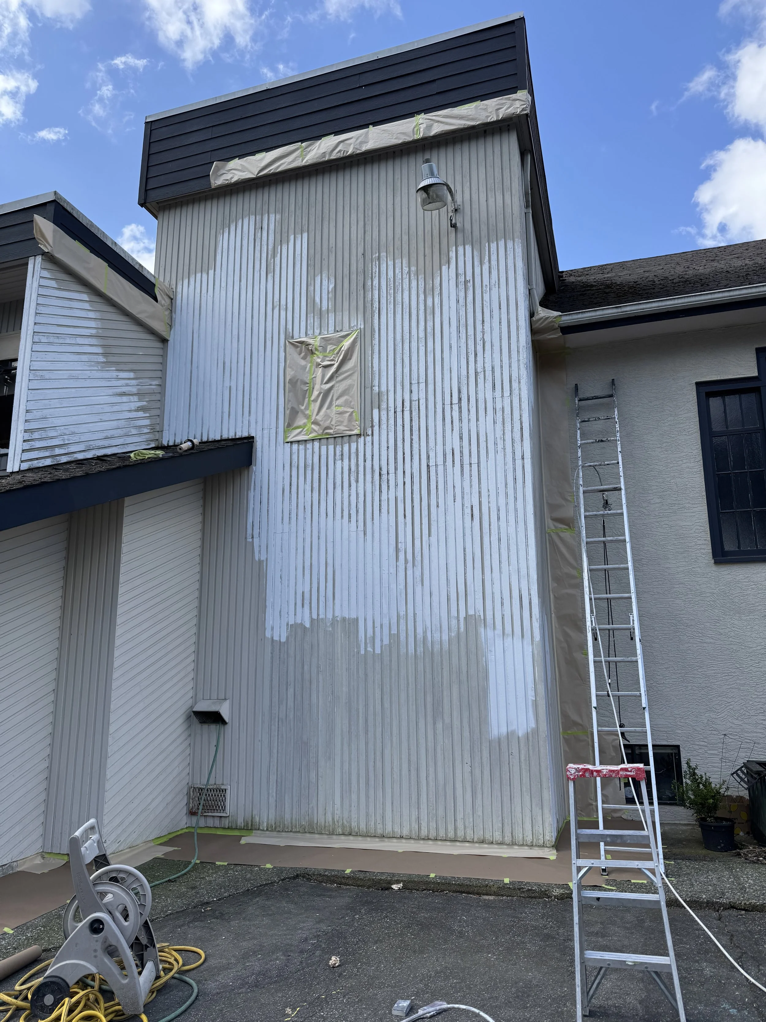 Exterior house wall being painted with white primer, with painter's tape on window and edges, ladder leaning against wall, and painting tools on ground.