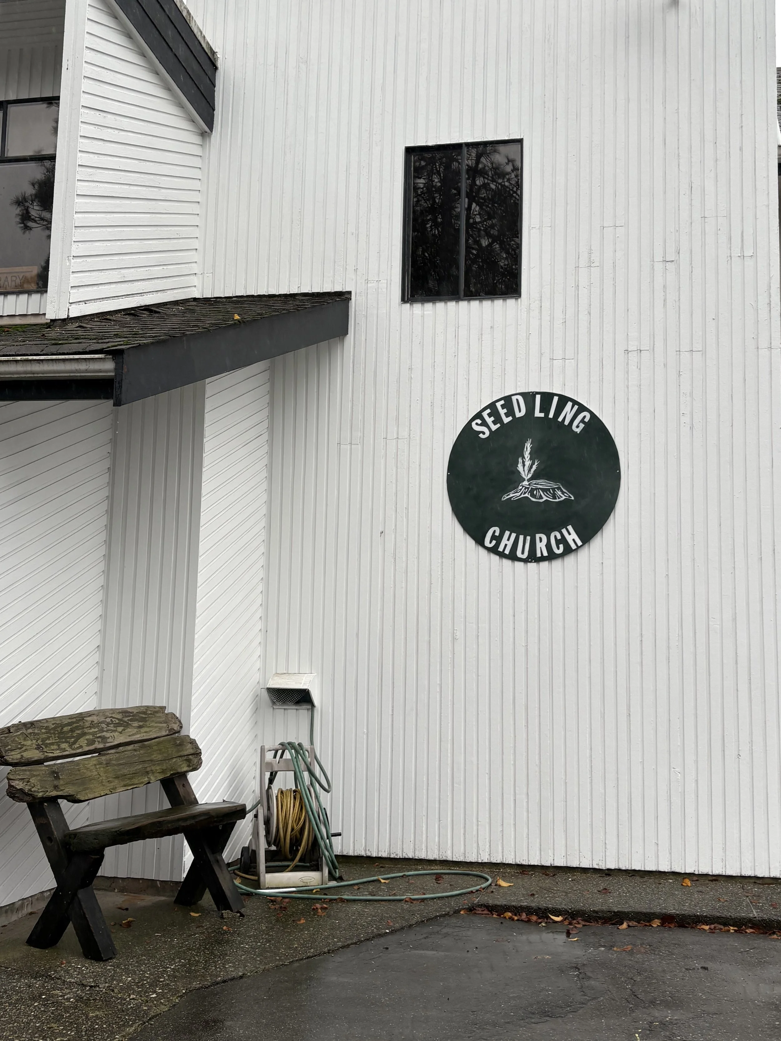 Exterior of Seed Ling Church with a round sign featuring a plant and mountain graphic, a weathered wooden bench, and garden hoses on a reel, on a wet concrete surface.