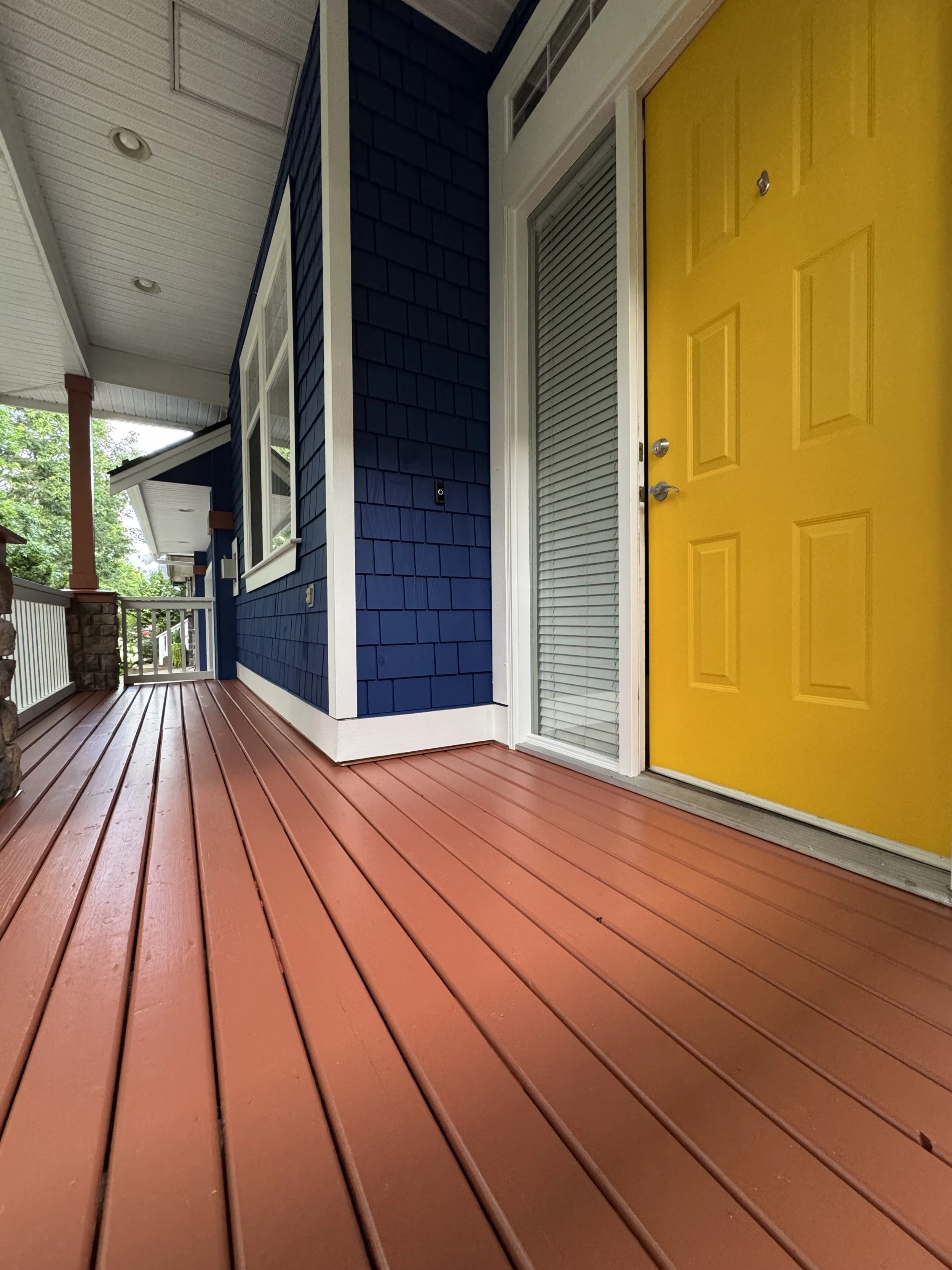 Colorful front porch with a mustard yellow door, blue siding, and reddish-brown wooden flooring.
