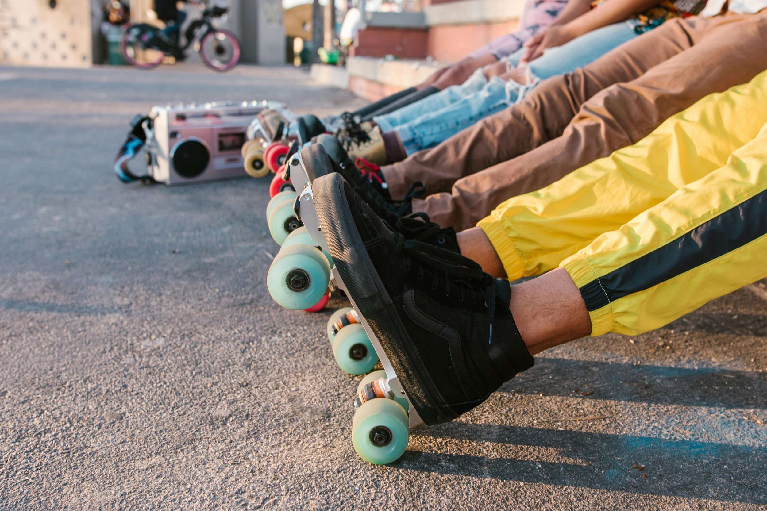 Six people sitting with legs extended, wearing roller skates, on an asphalt surface. In the background, there is a boombox and pink bicycle.