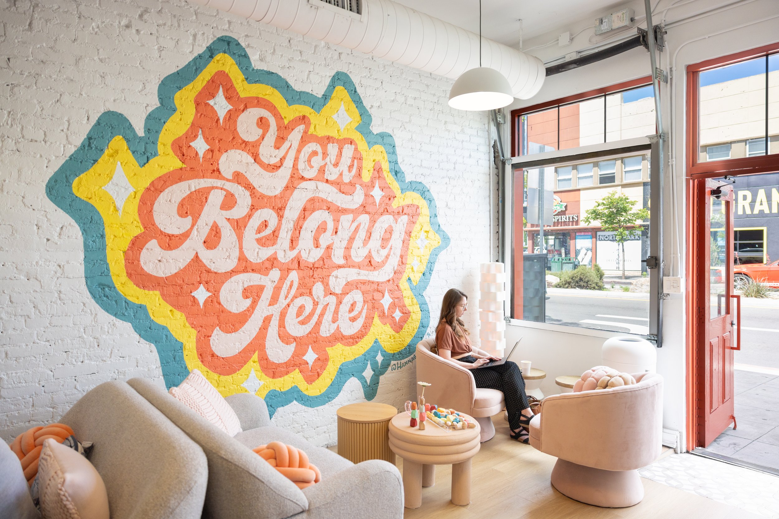 Interior of a bright, modern shop or cafe with white brick walls, a large colorful mural reading "You Got This Here," and large windows revealing a street scene outside. There are cozy seating areas with sofas, chairs, and small tables, and a woman is sitting with a laptop.